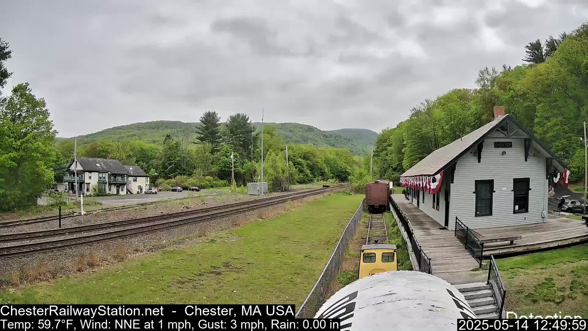 A small yellow train approaches a white train station on a slightly overcast day, with green hills and trees in the background.