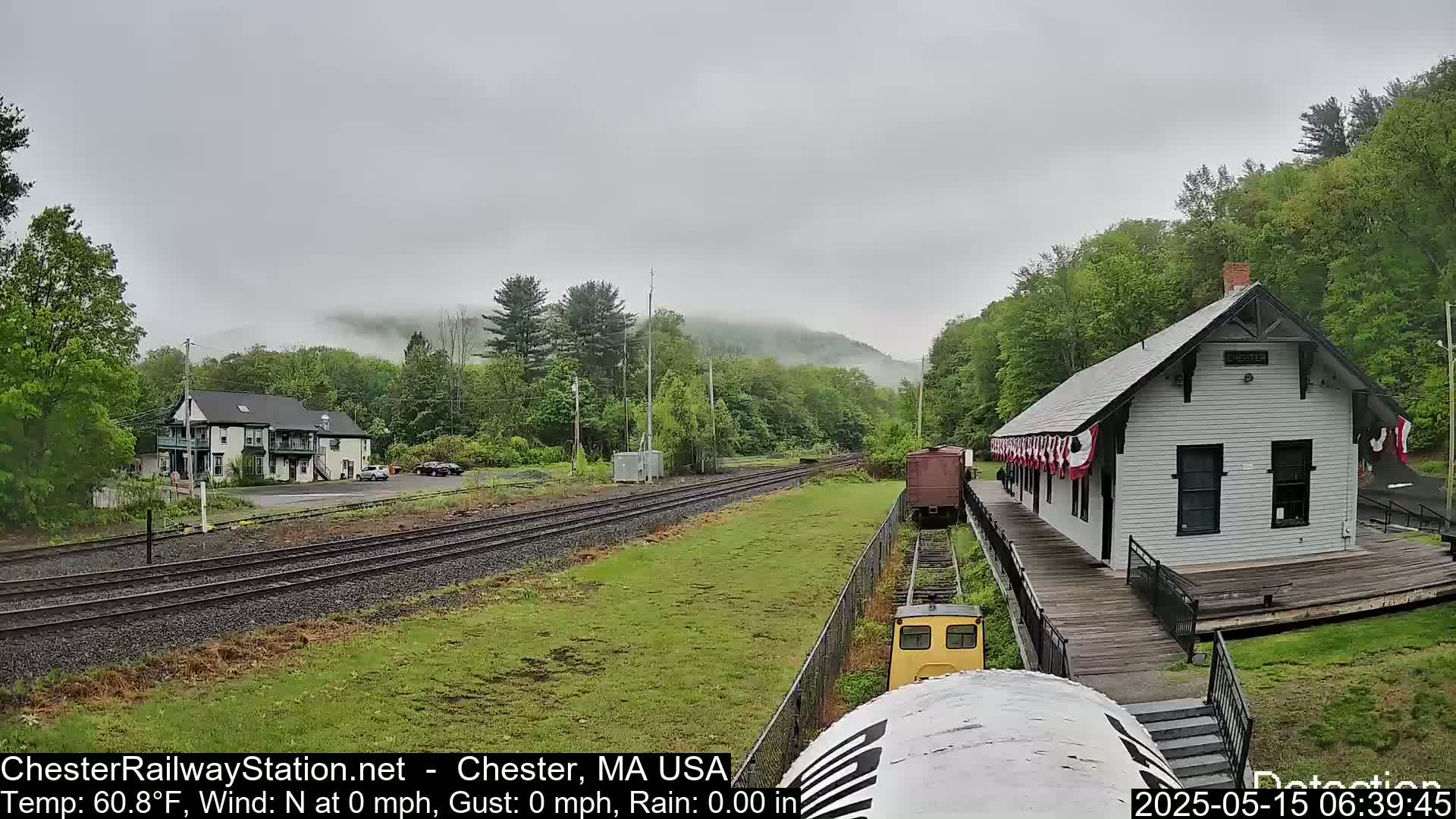A train approaches a white railway station nestled among green trees and hills shrouded in fog under an overcast sky.