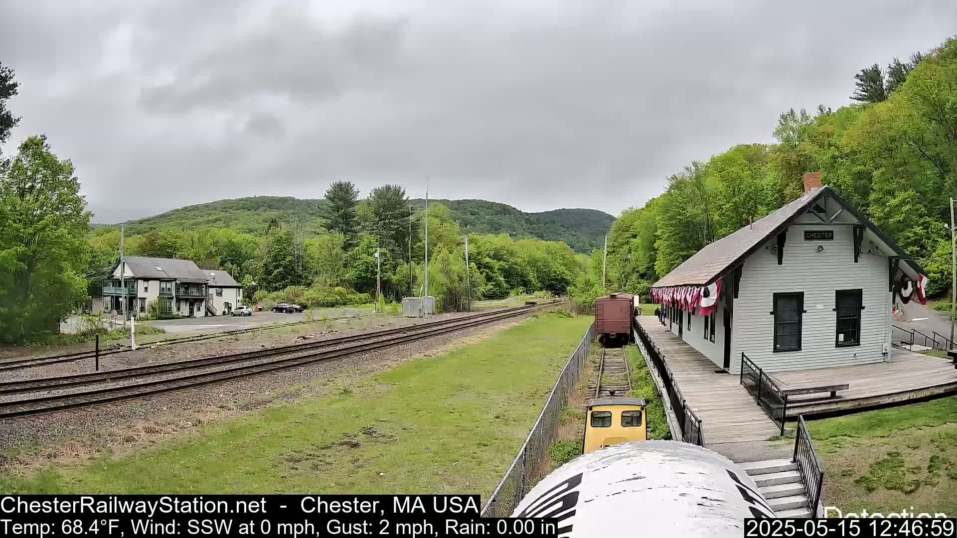 A train approaches a small, white train station nestled in a verdant valley under an overcast sky.