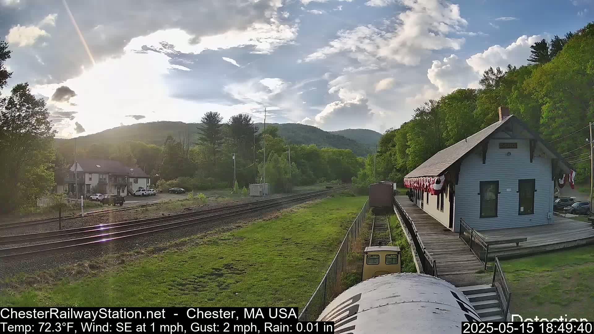 A train approaches a small, white train station nestled in a verdant valley under an overcast sky.