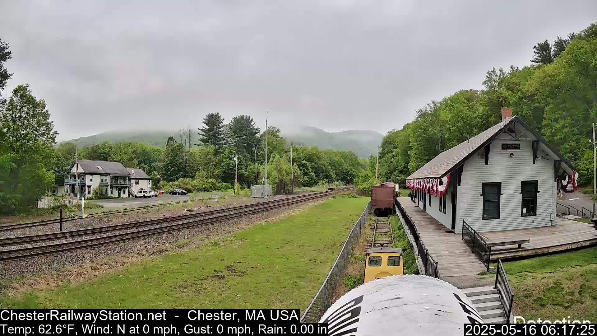 A small train approaches a white train station nestled among green trees and hills under an overcast sky.