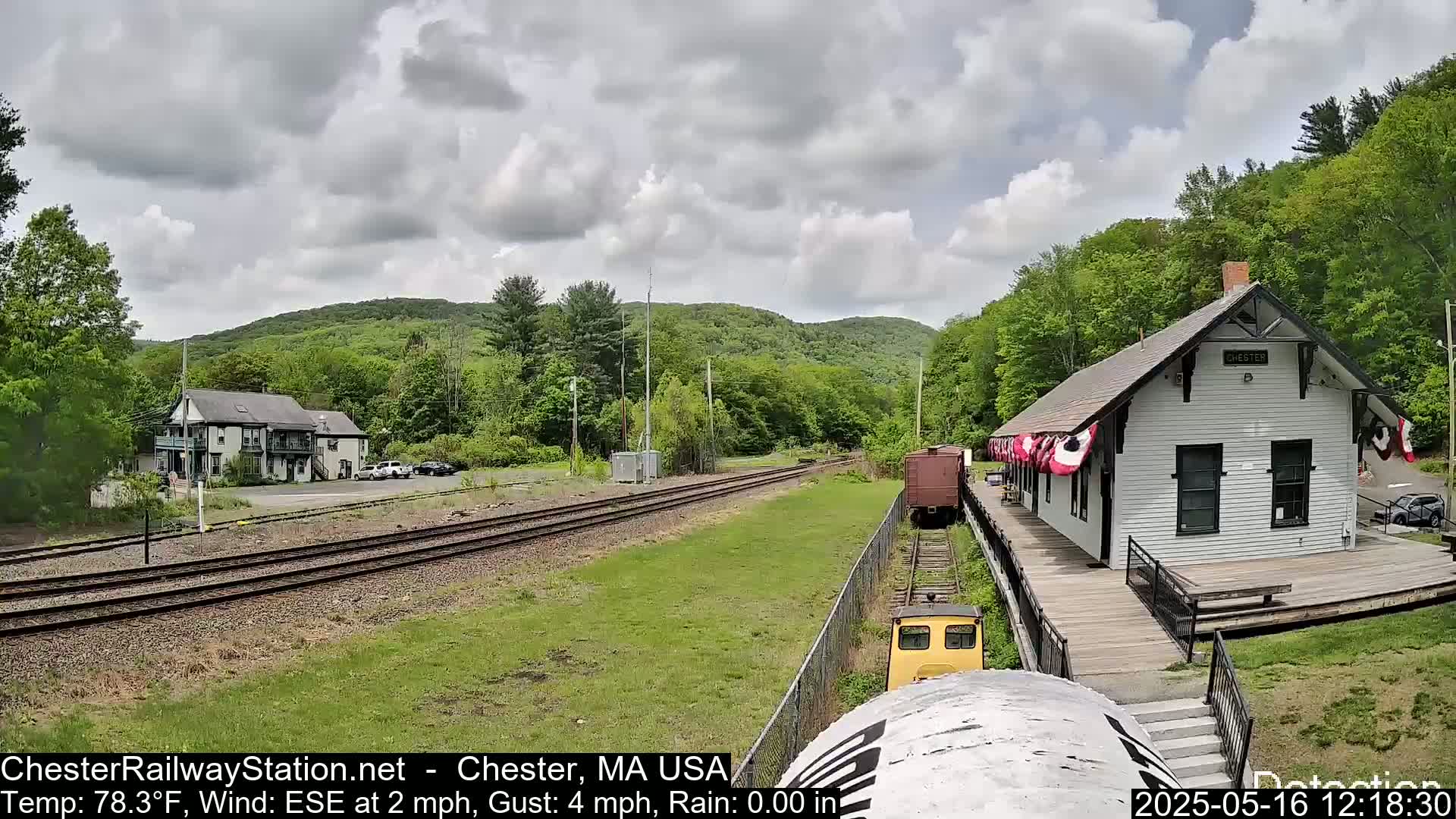 A train approaches a white train station on a partly cloudy day, with green hills and trees in the background.
