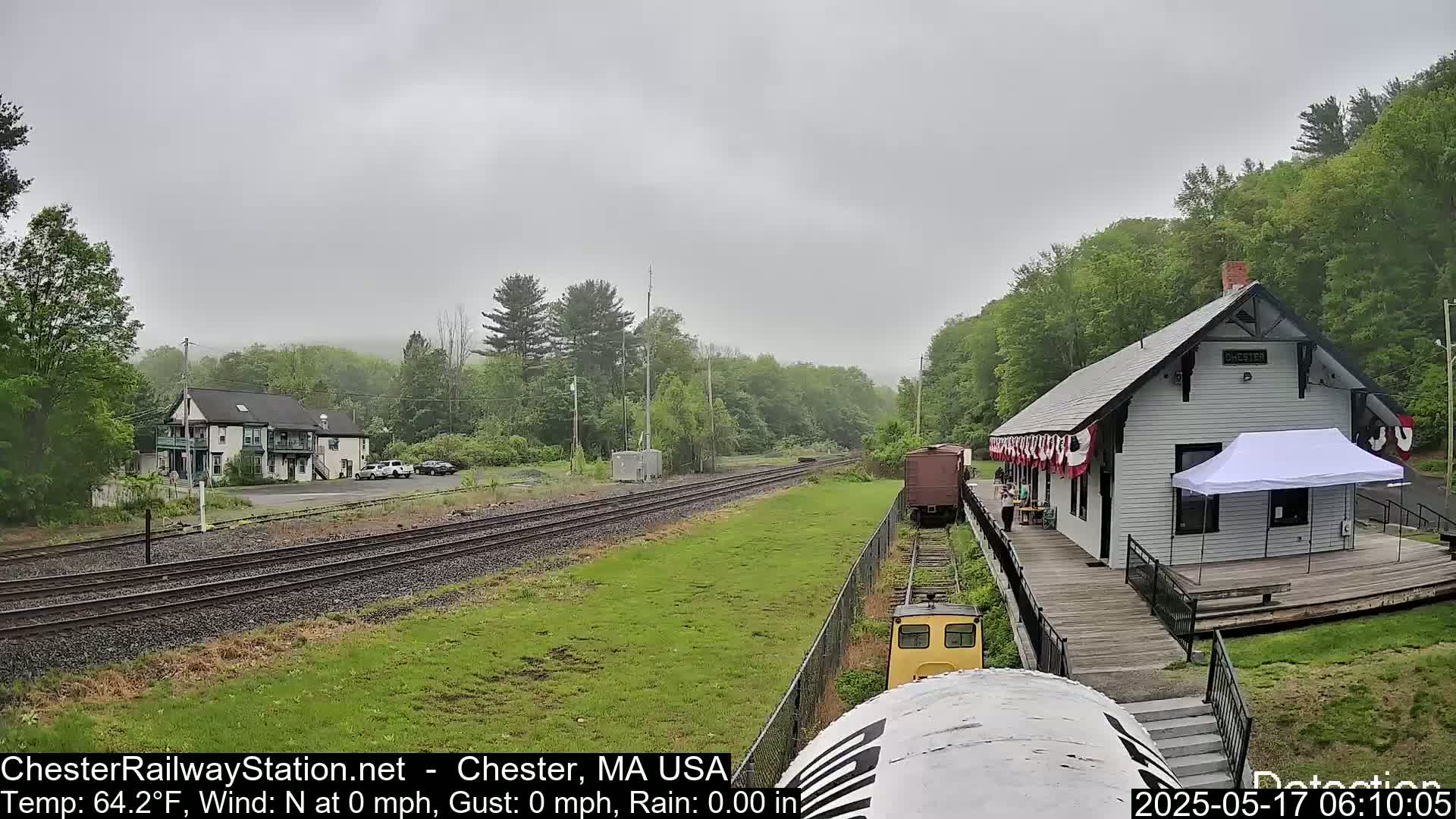 A train approaches a small, white train station situated amongst lush green trees on an overcast day.