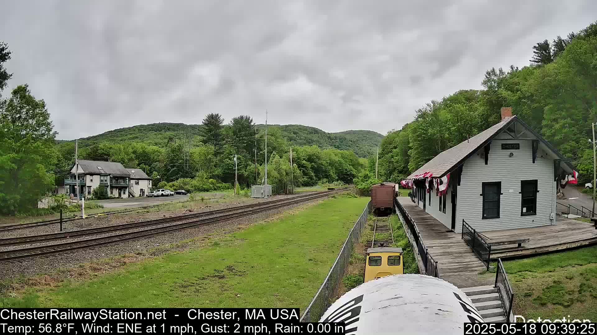 A train approaches a small, white train station nestled in a green, hilly landscape under an overcast sky.