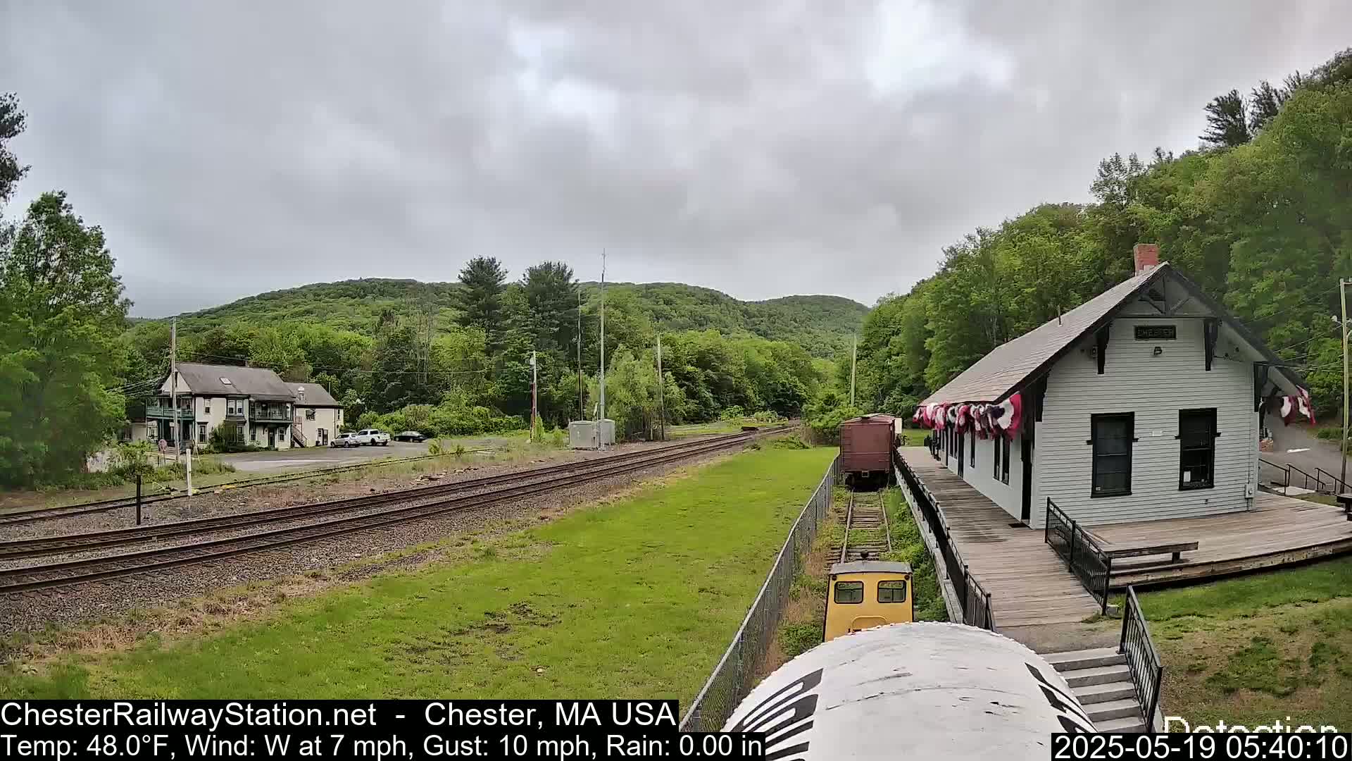 A small train approaches a white train station situated on a grassy area next to railroad tracks, with a cloudy sky and green hills in the background.