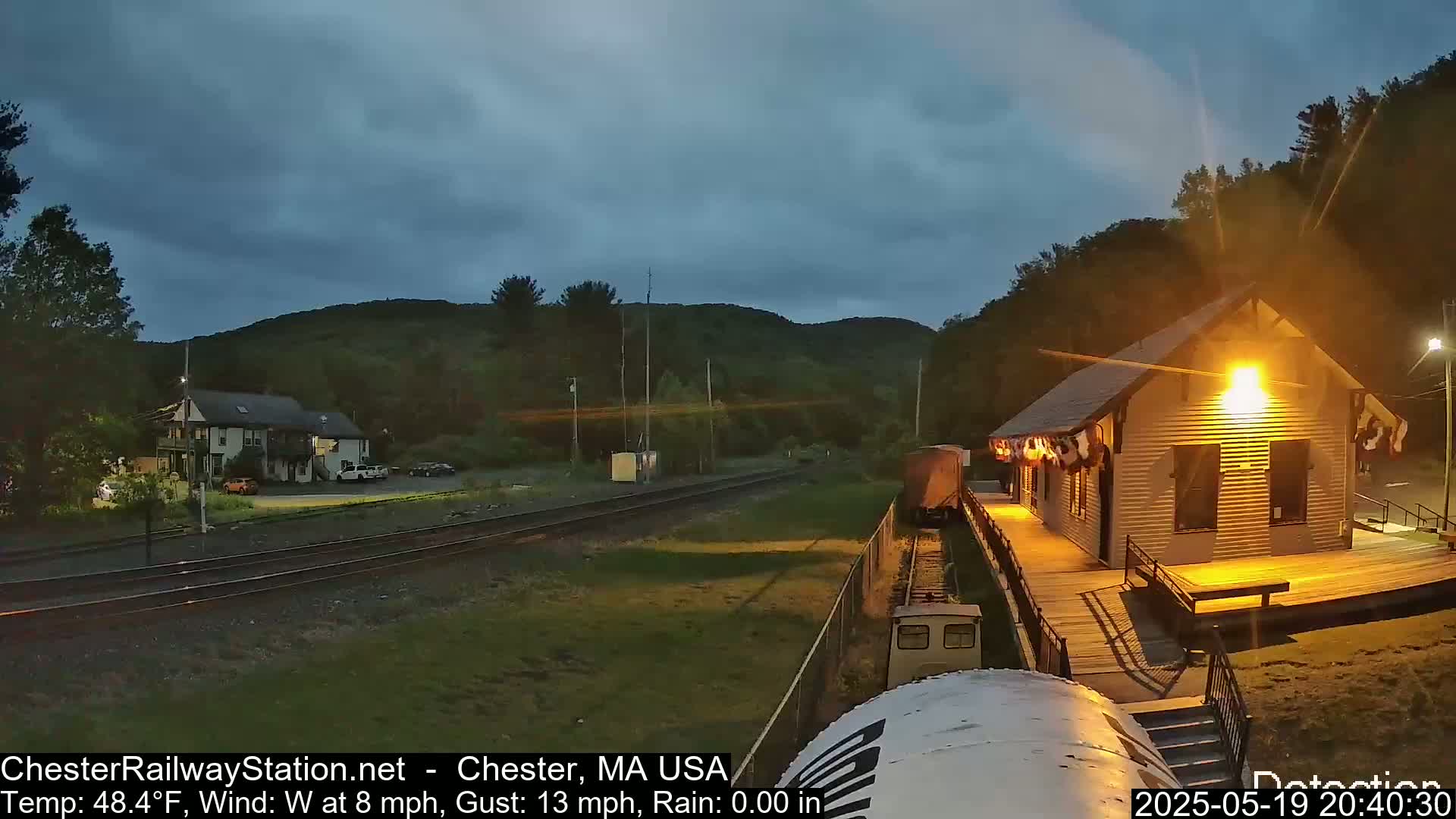 A train approaches a small, lit station at dusk under a partly cloudy sky.