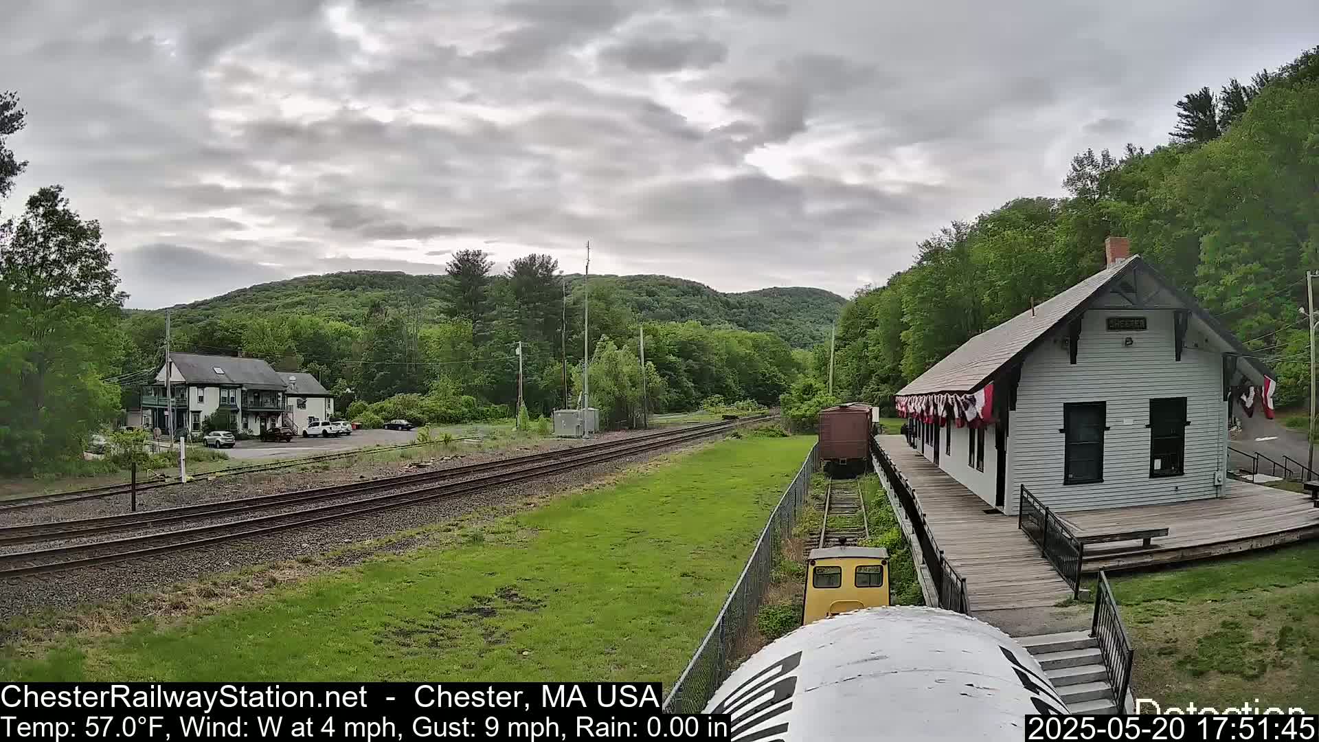 A small train approaches a white train station on a partly cloudy day, with a grassy area and a wooded hillside in the background.