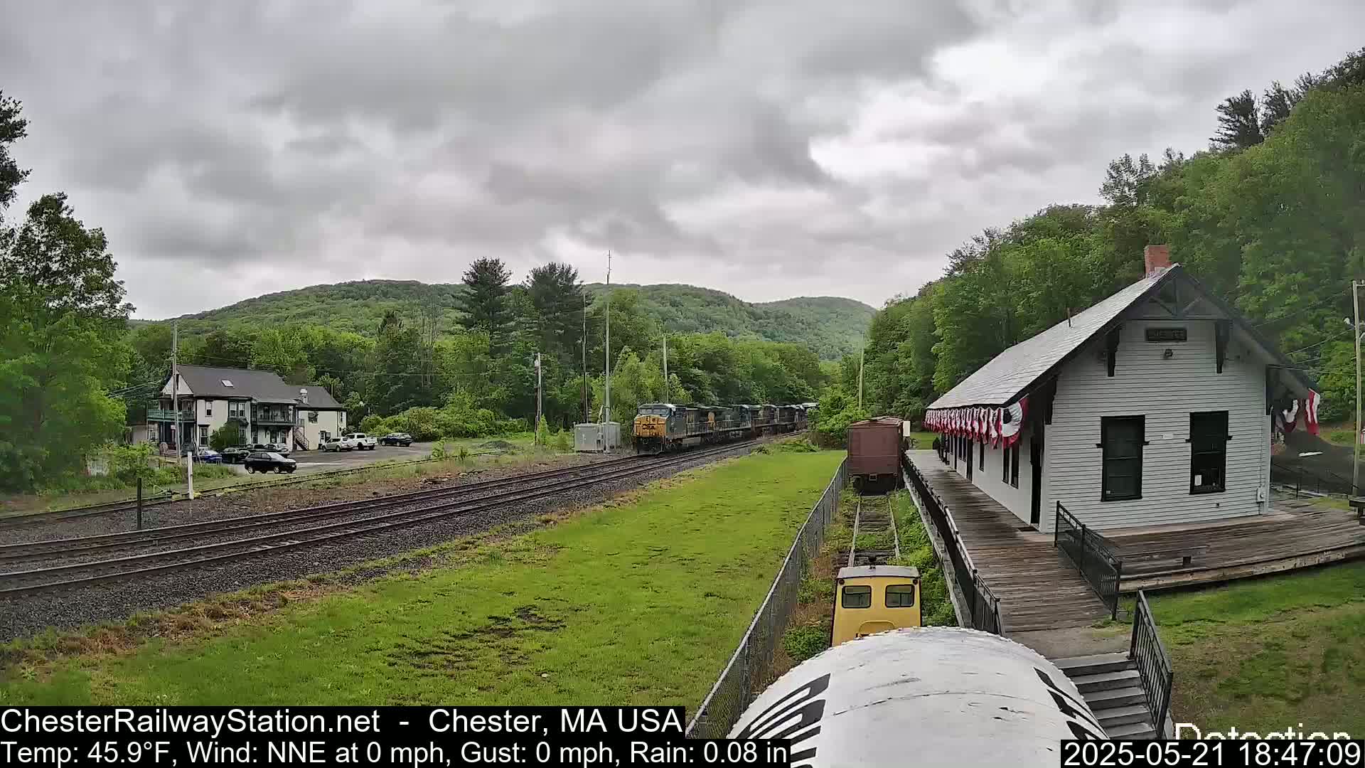 A freight train passes a small white train station on a rainy day, with green hills and trees in the background.
