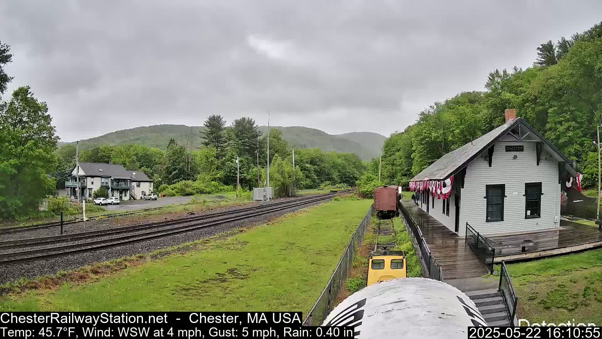 A view of a train station and railroad tracks on a rainy day, with green hills and trees in the background.