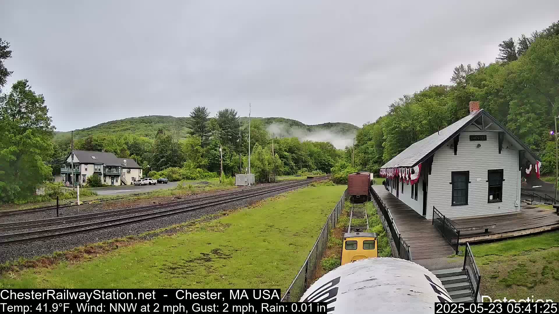 A small train approaches a white train station beside a two-story building, all under a cloudy, rainy sky.