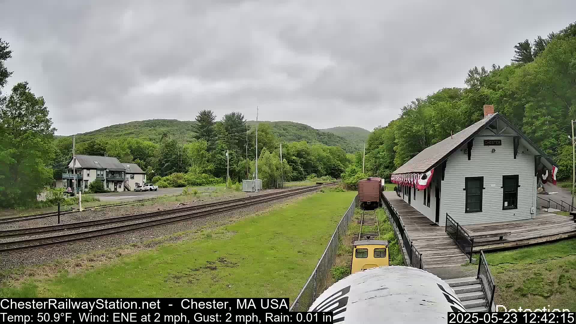 A view of a train station and tracks on a lightly rainy day, with a small building and hills in the background.