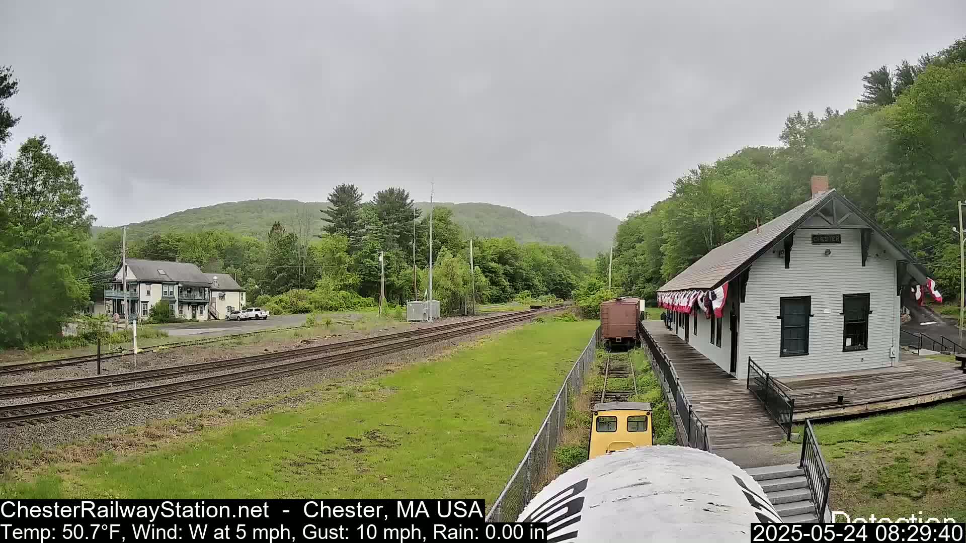 A train station sits beside railroad tracks in a verdant valley under an overcast sky.