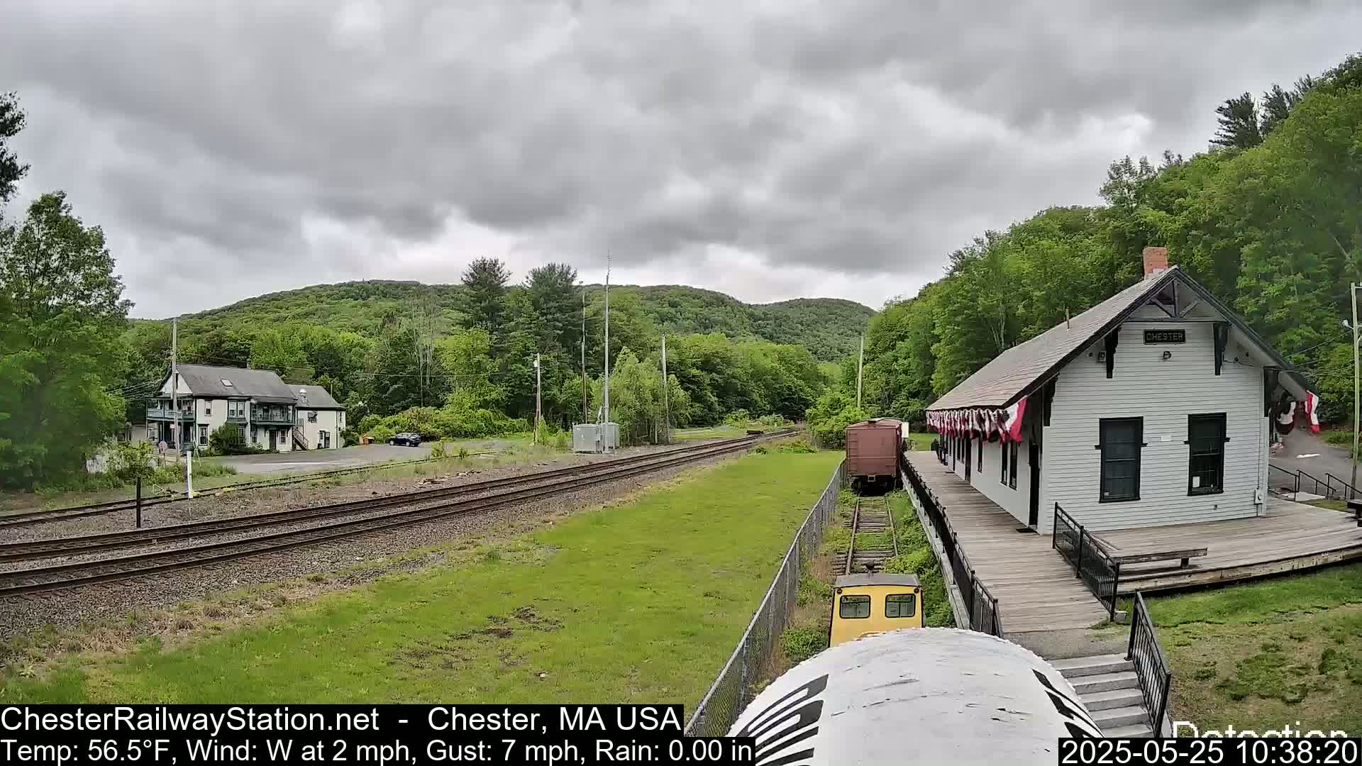 A small yellow train approaches a white train station next to railroad tracks, under a cloudy sky, situated in a green, hilly landscape.