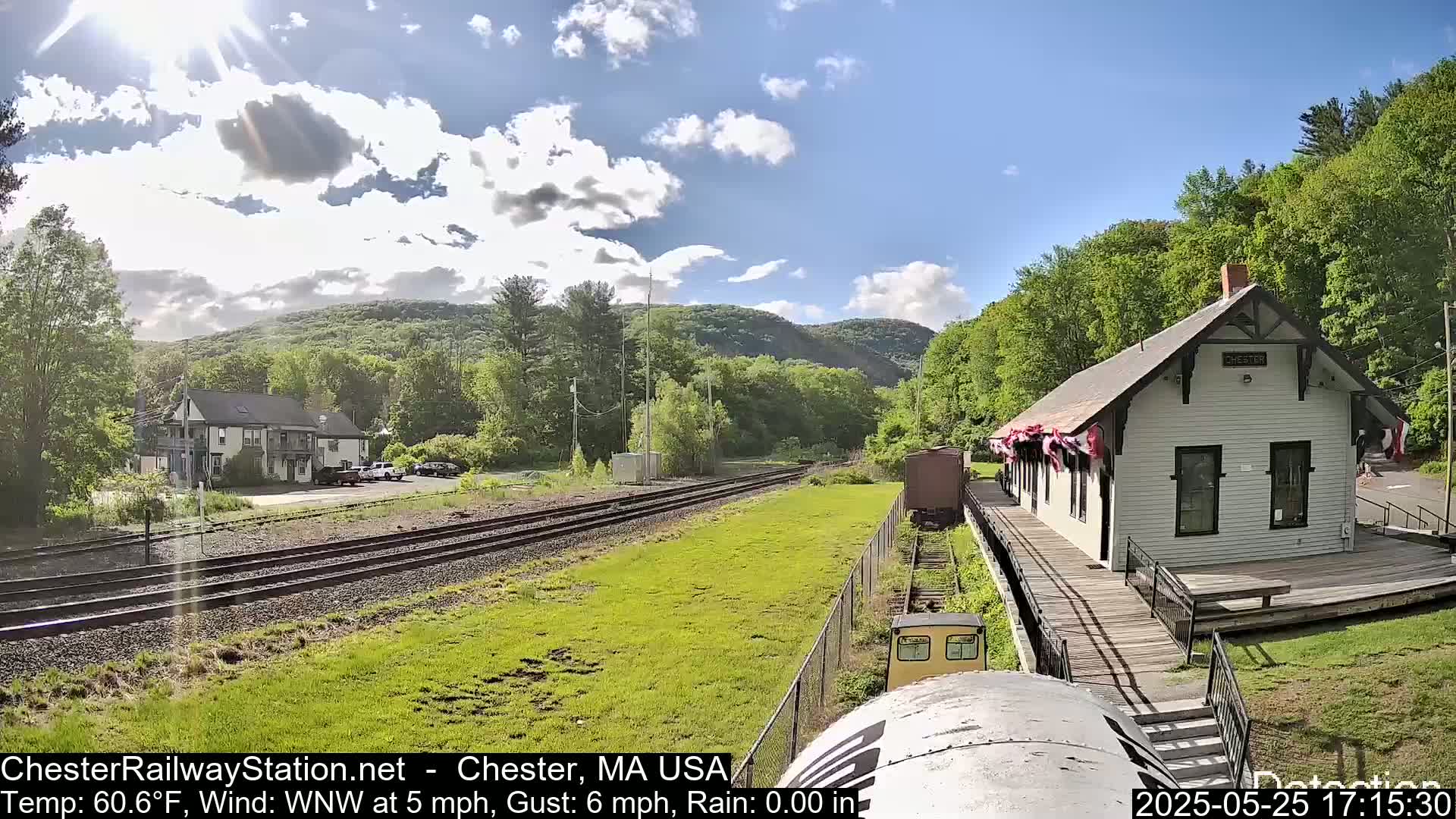 A sunny day with partly cloudy skies shows a train approaching a white train station next to railroad tracks, with a building and green hills in the background.