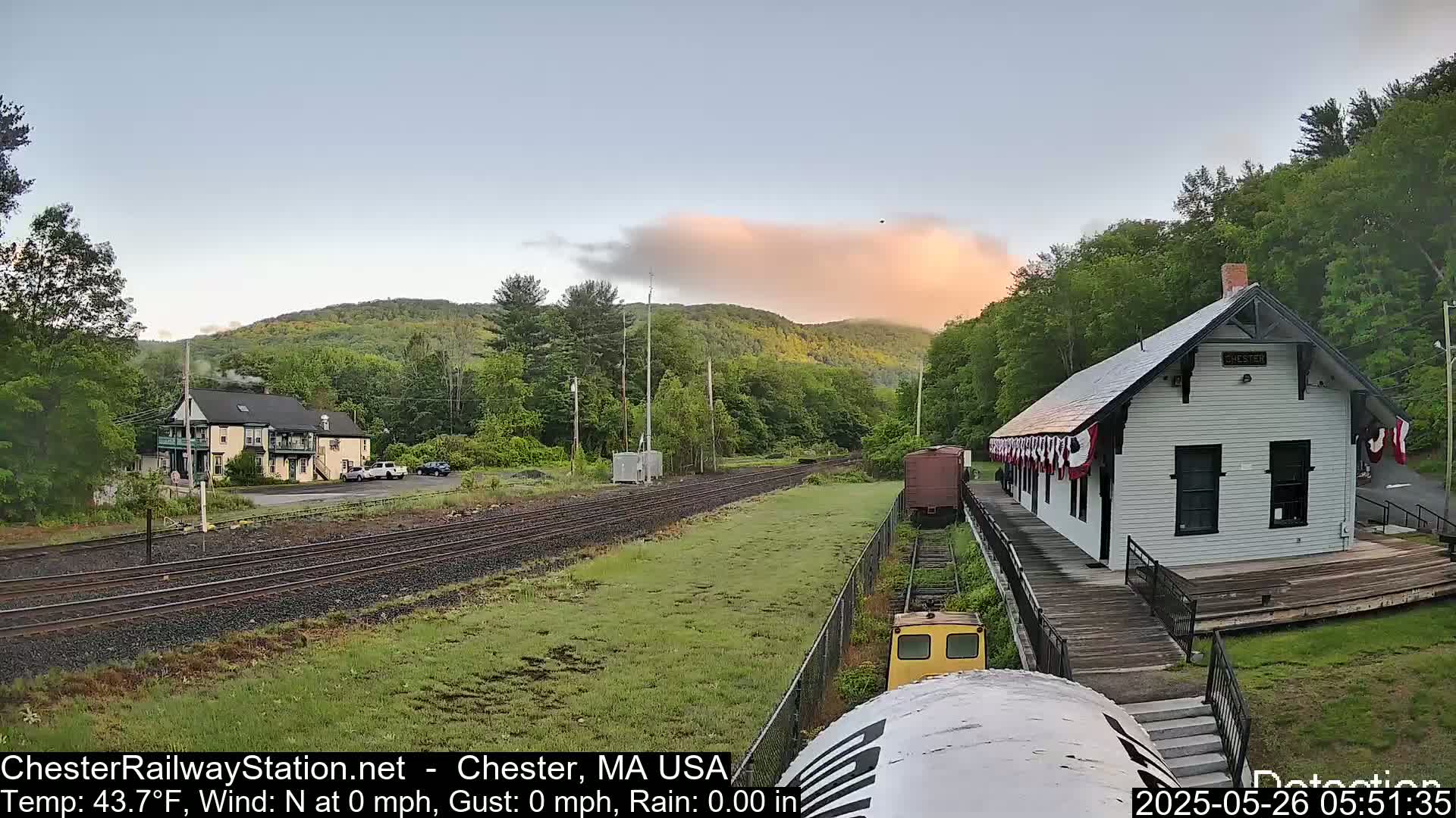 A small train approaches a white train station decorated with flags, situated on a grassy area next to train tracks with a hillside in the background under a partly cloudy, sunrise sky.