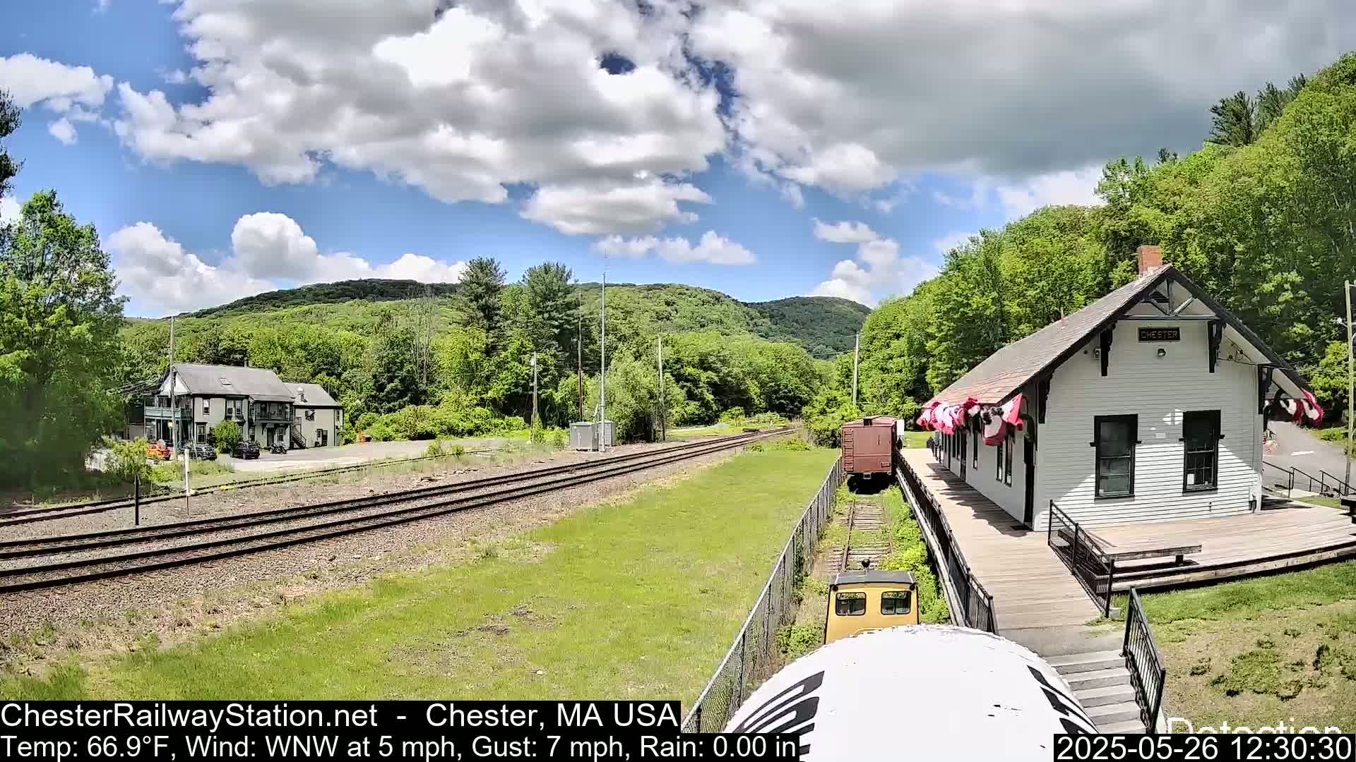 A partly cloudy day shows a train station, train cars, and tracks next to a building and forested hills.