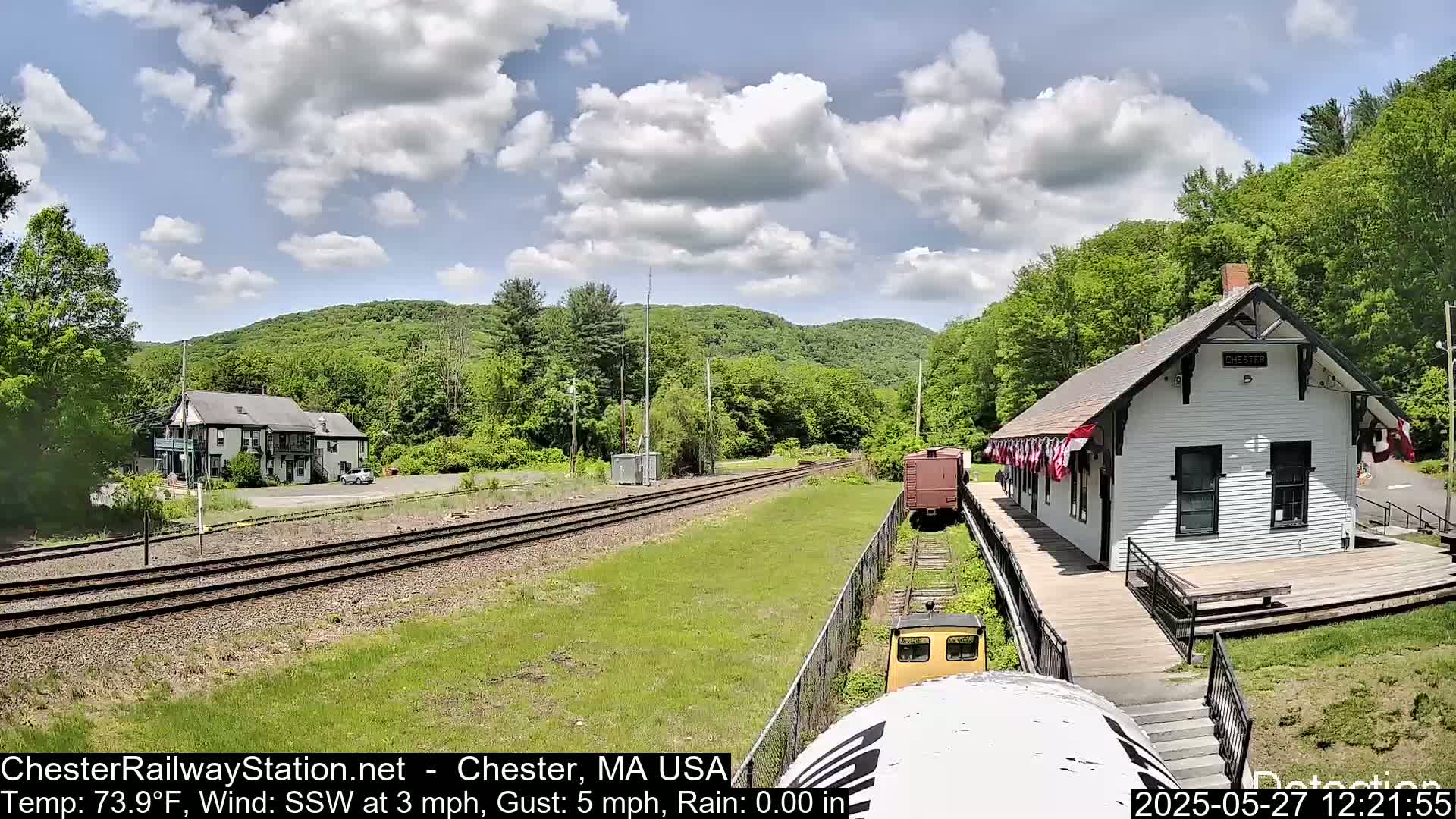 A train approaches a small, white train station nestled in a verdant valley under a partly cloudy sky.