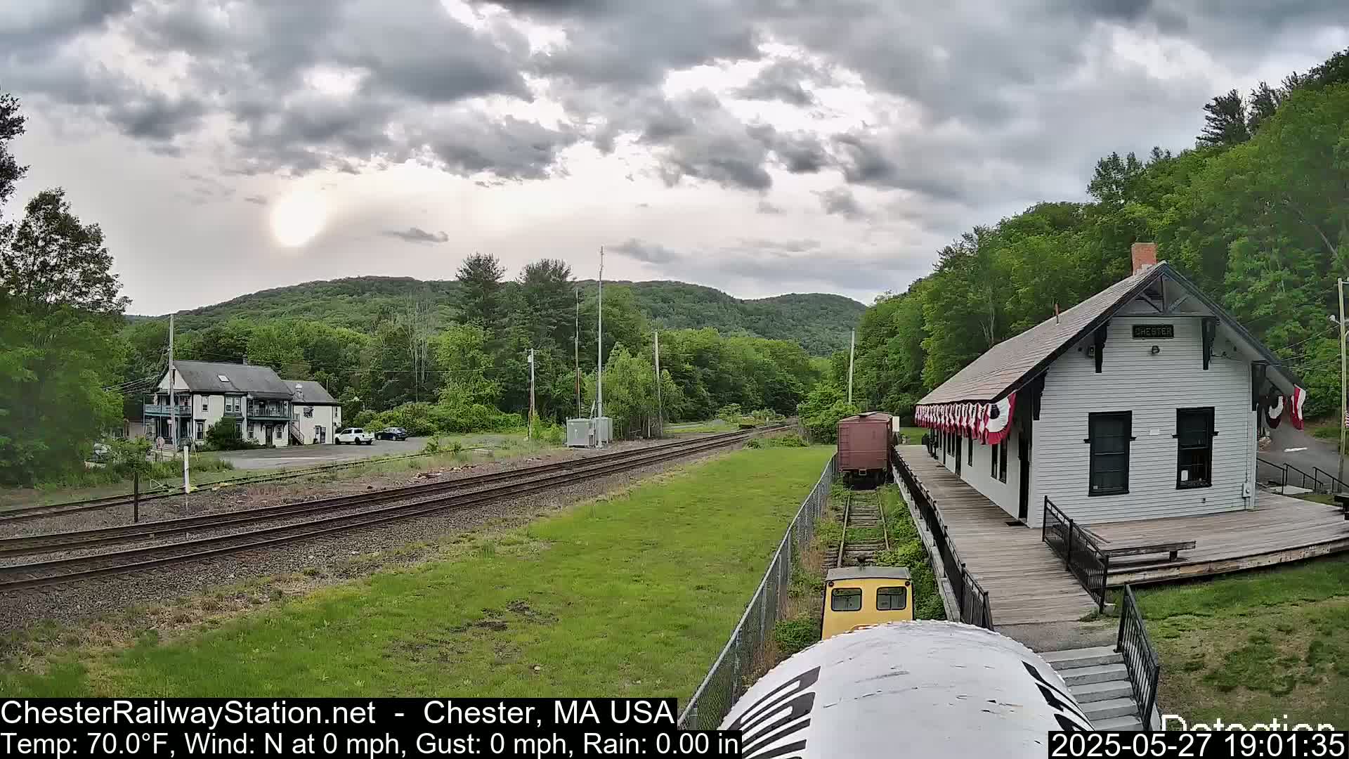 A small train approaches a white train station on a mostly cloudy day, with green hills and trees in the background.
