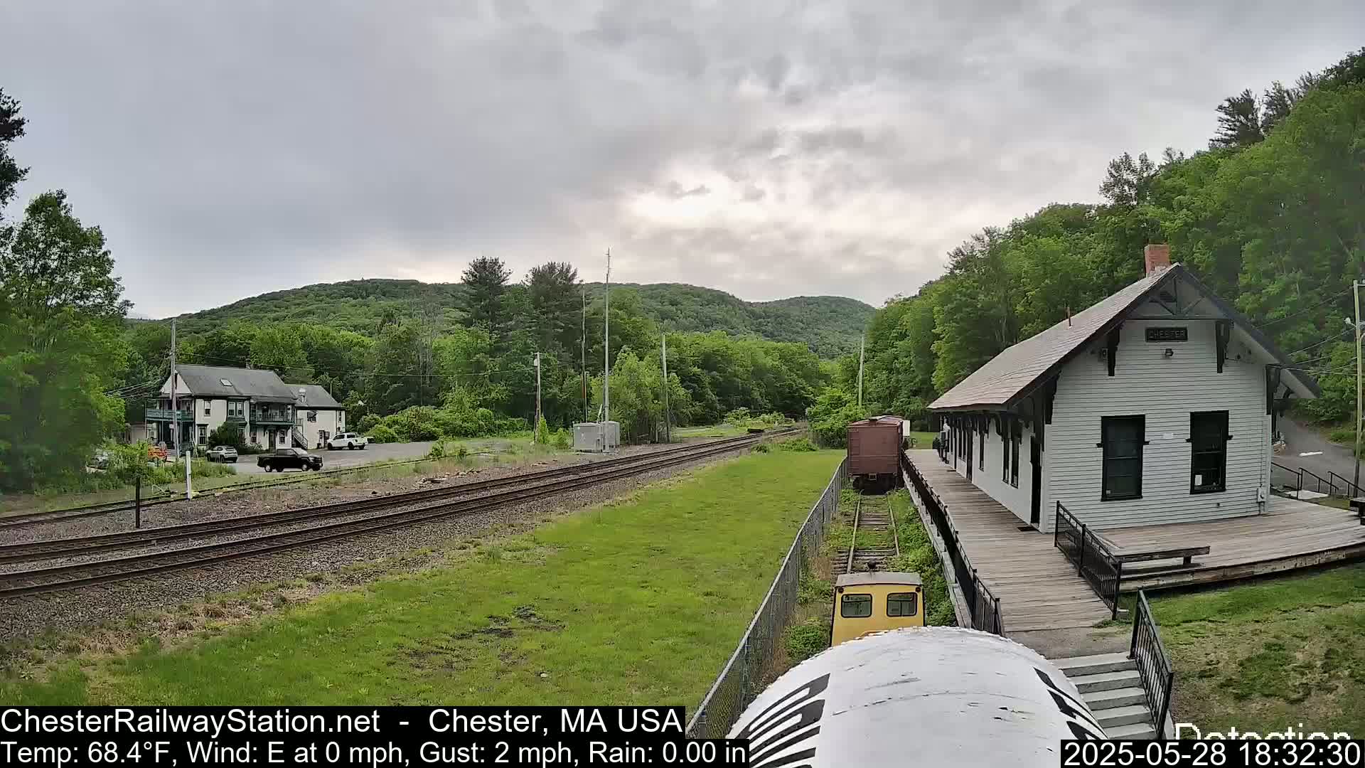 A train approaches a white train station situated next to a two-story building, all surrounded by lush green trees and hills under an overcast sky.