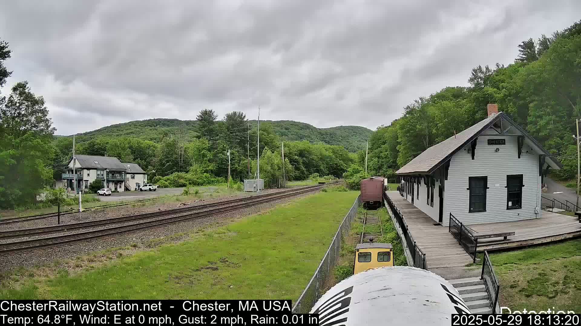 A small, white train station sits beside railroad tracks on a slightly overcast day, with light rain and lush green hills in the background.
