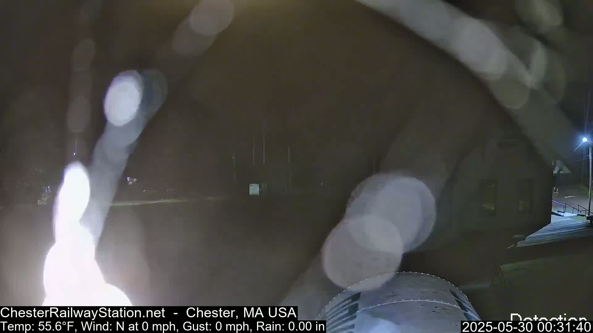A nighttime, rainy view shows blurry lights reflecting on a rain-covered camera lens, partially obscuring a dark building and surrounding area.