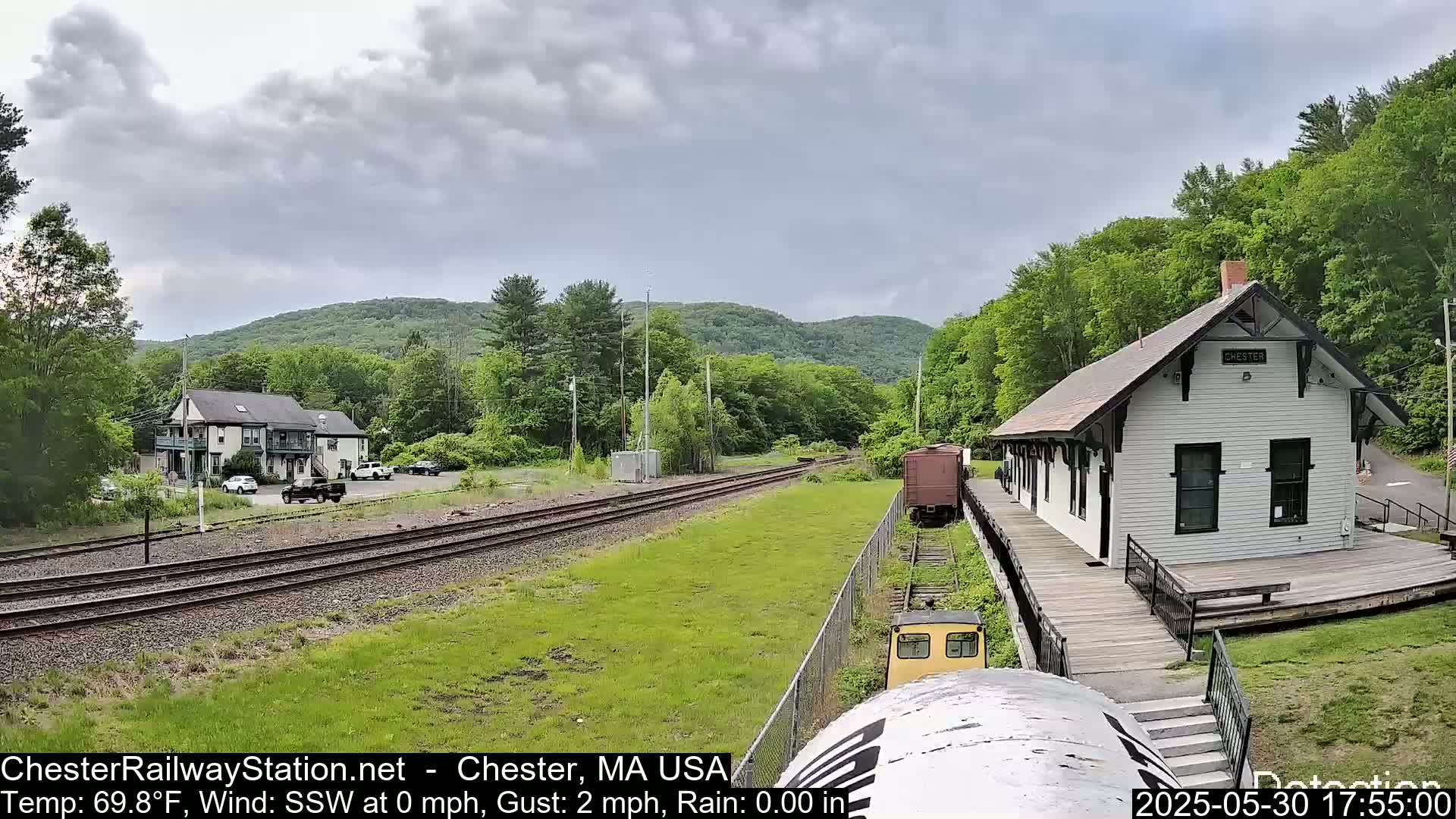 A train approaches a white train station situated beside a grassy area, with a cloudy sky overhead and forested hills in the background.