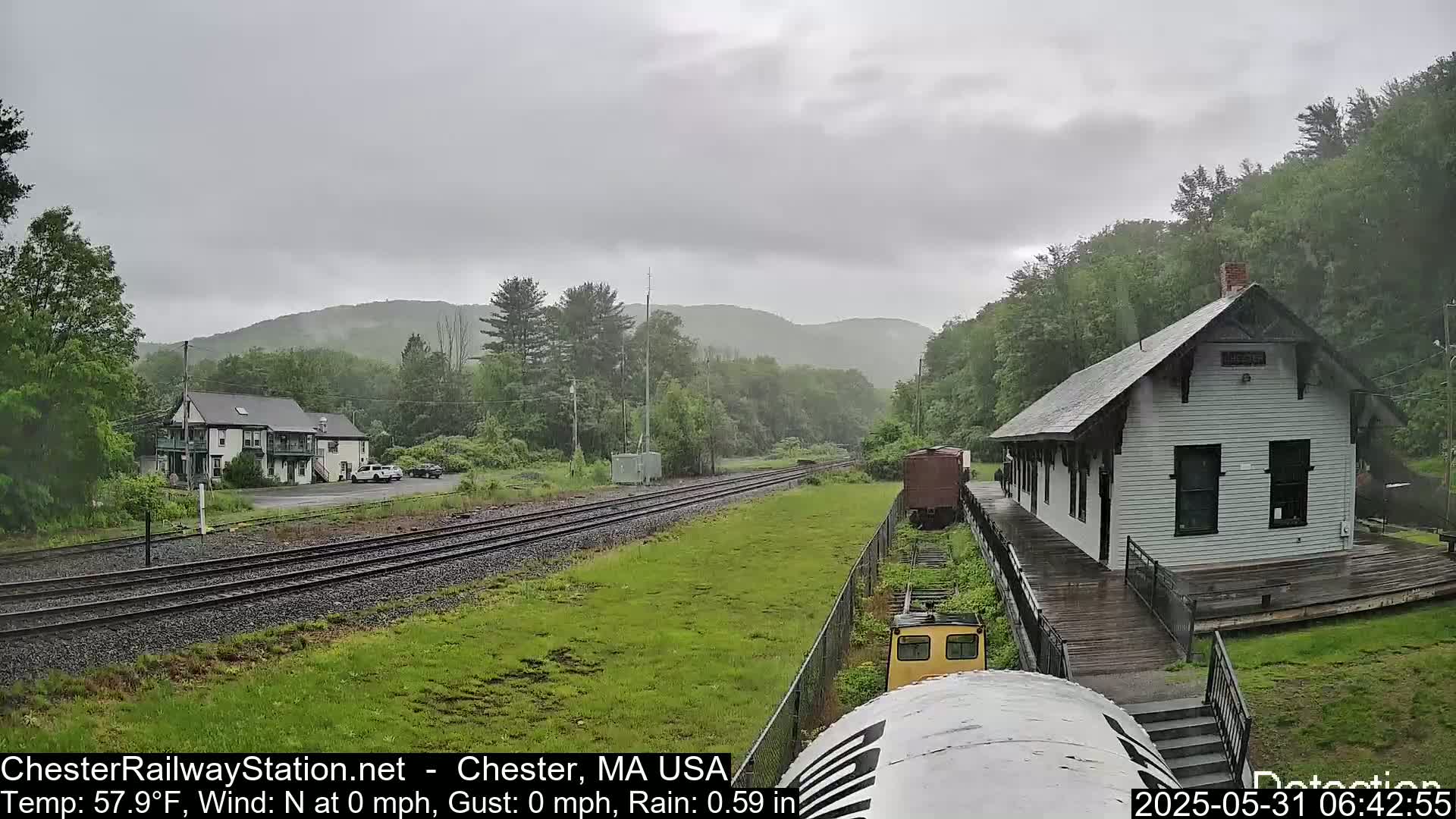 A rain-soaked train station sits beside train tracks, with a two-story building and green hills visible in the background under a gray sky.