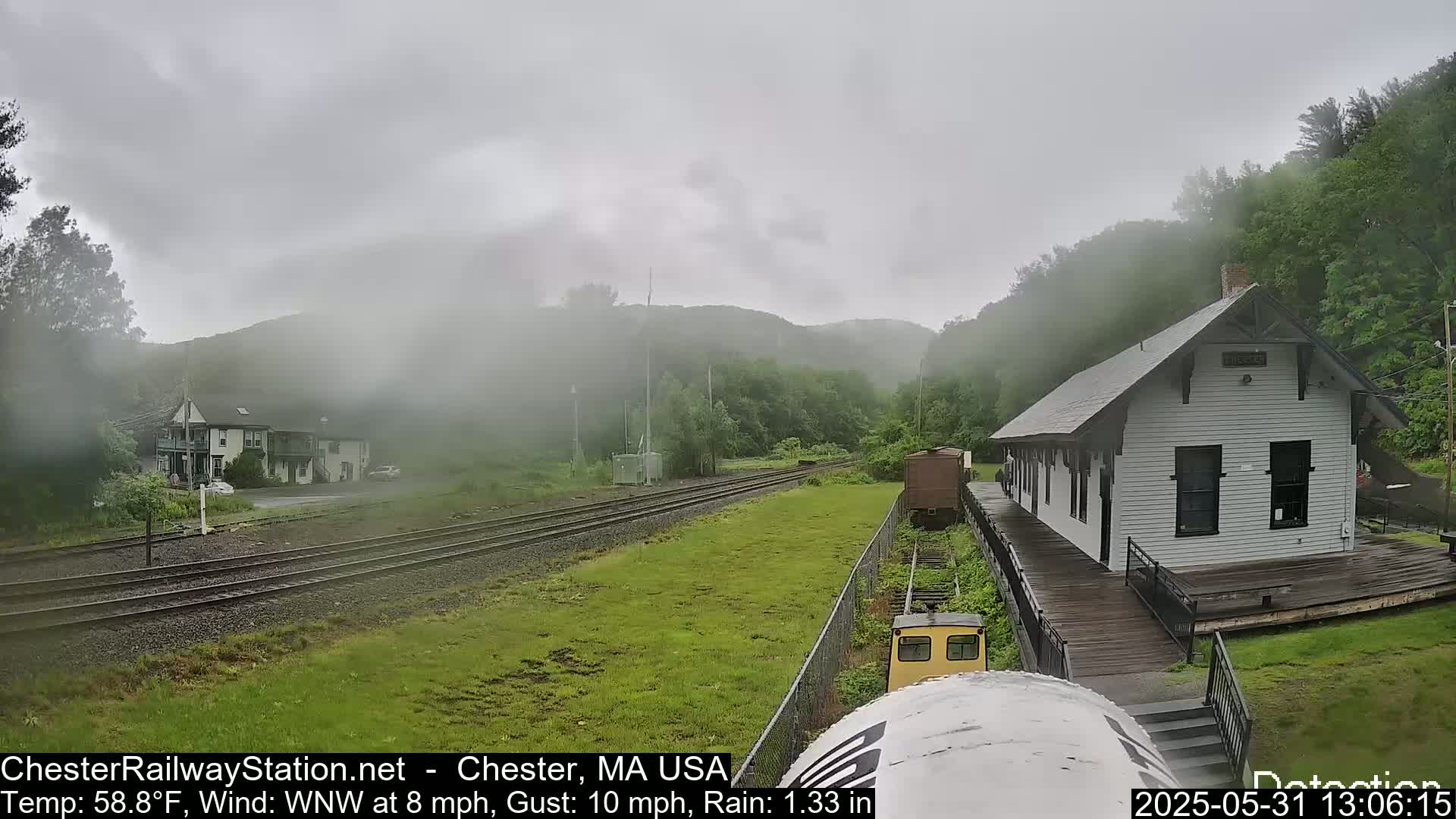 A train depot sits beside railroad tracks on a rainy day, with misty hills and greenery in the background.