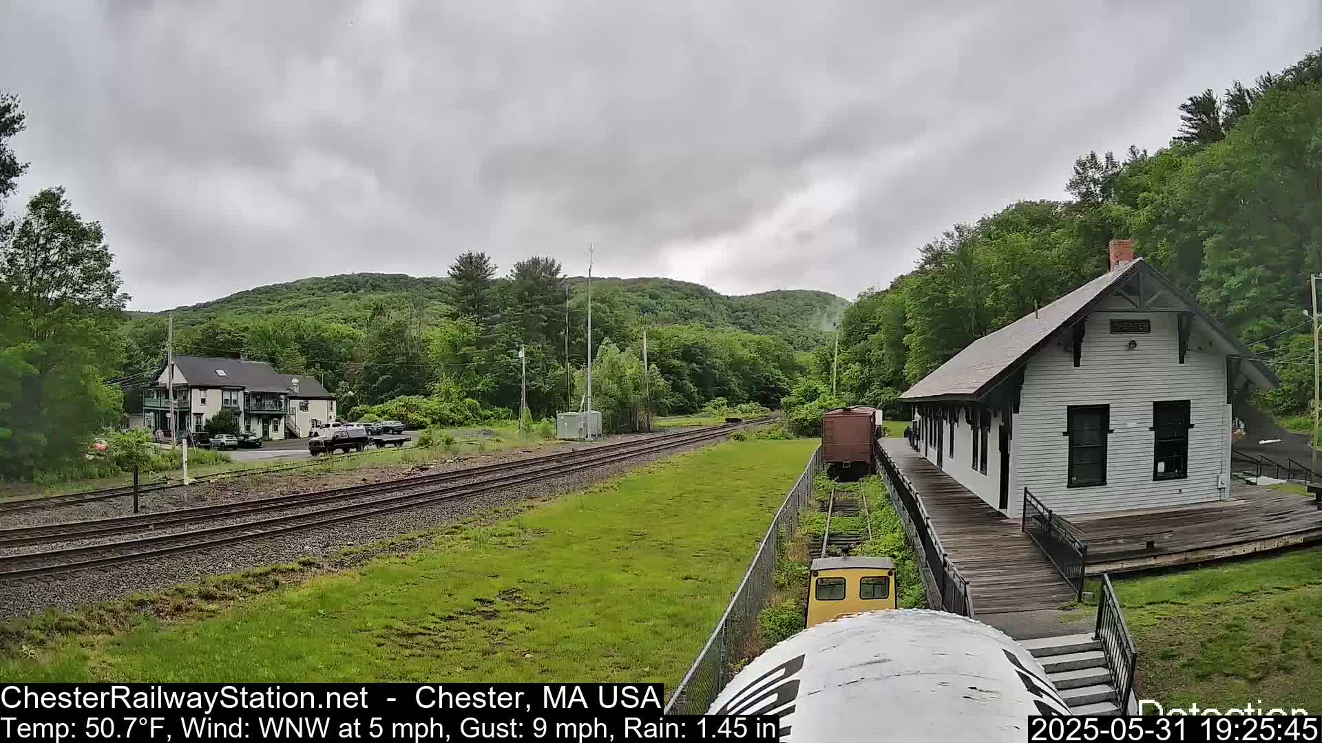 A small train approaches a white train station nestled in a green, hilly landscape under a cloudy, rainy sky.