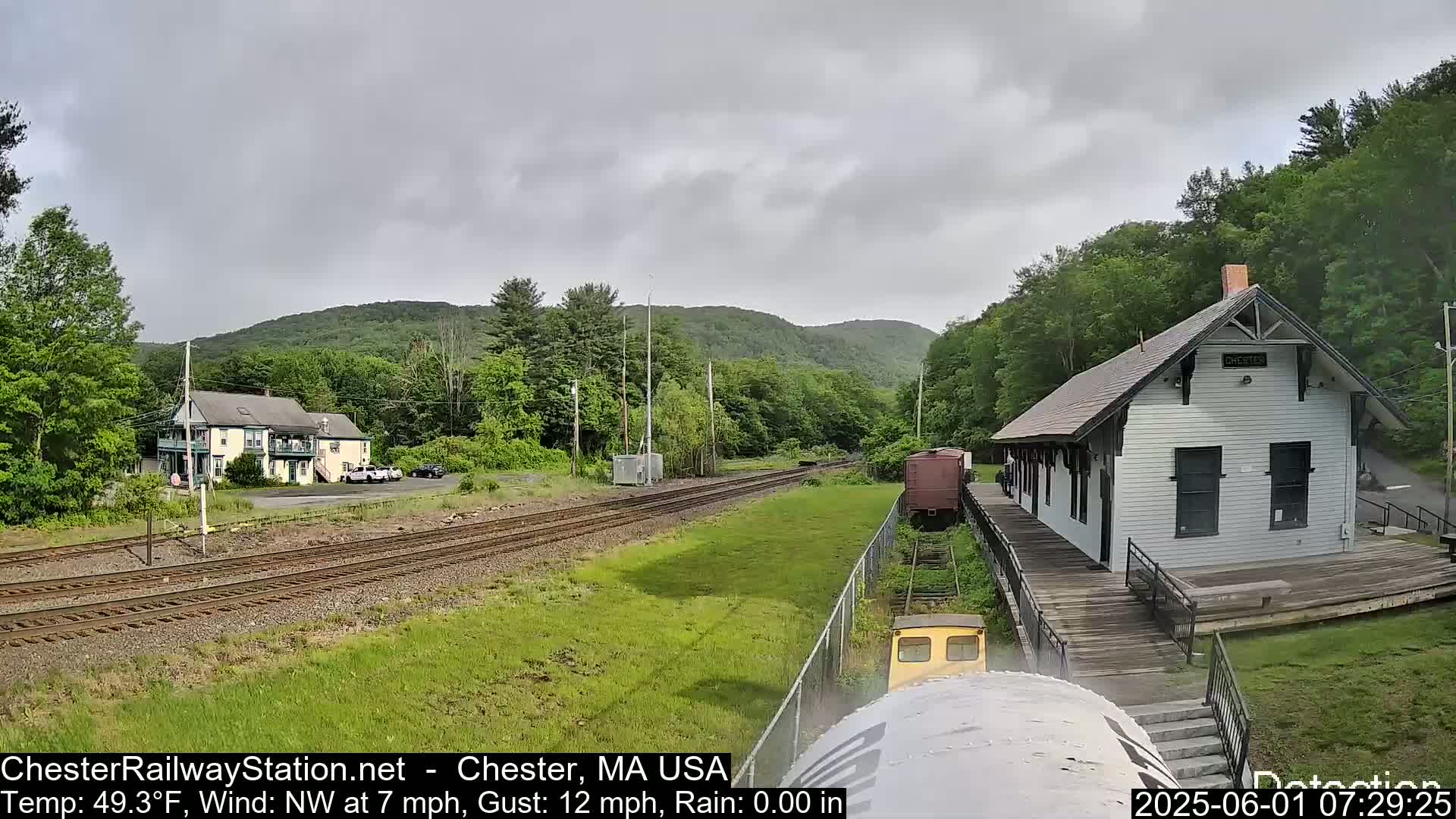 A train approaches a small, white train station situated beside train tracks and green hills under an overcast sky.