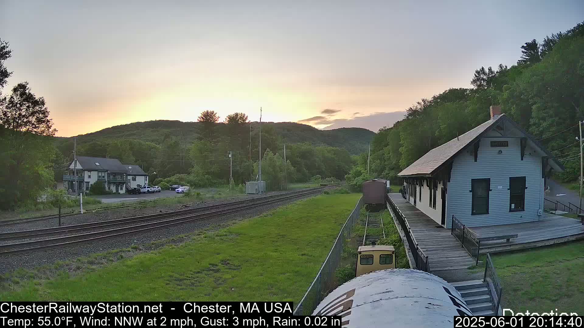 A train station and a two-story building are visible at sunset, with a grassy area and railroad tracks in between them, under partly cloudy skies.