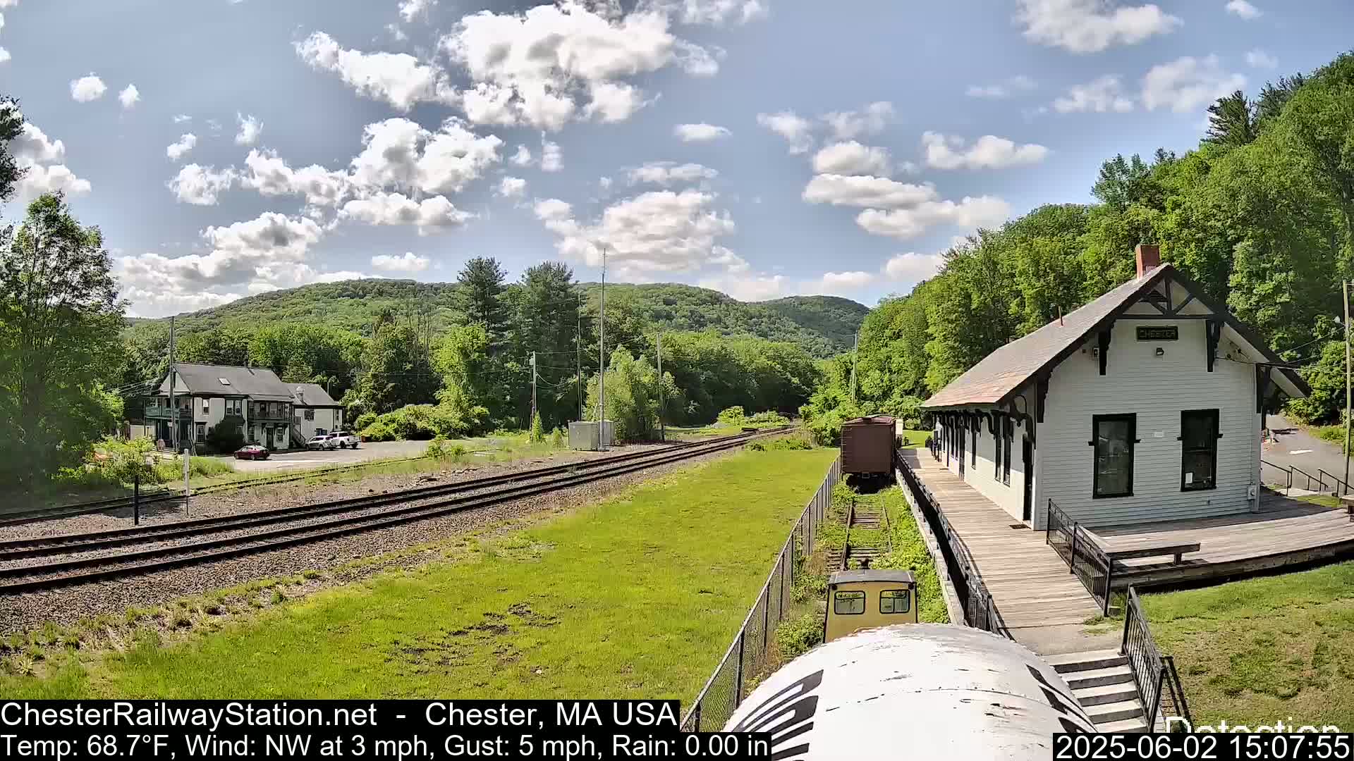 A partly cloudy day shows a train approaching a white train station next to a two-story building, all surrounded by green hills and trees.