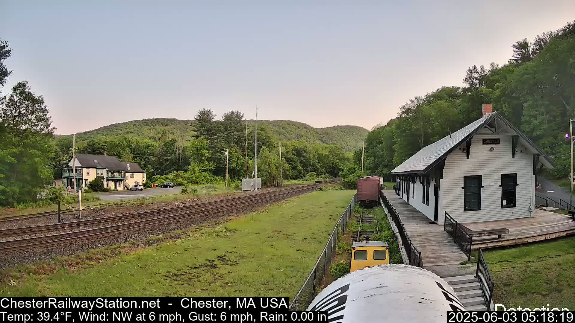 A train approaches a small, white train station nestled among green hills on a partly cloudy day.