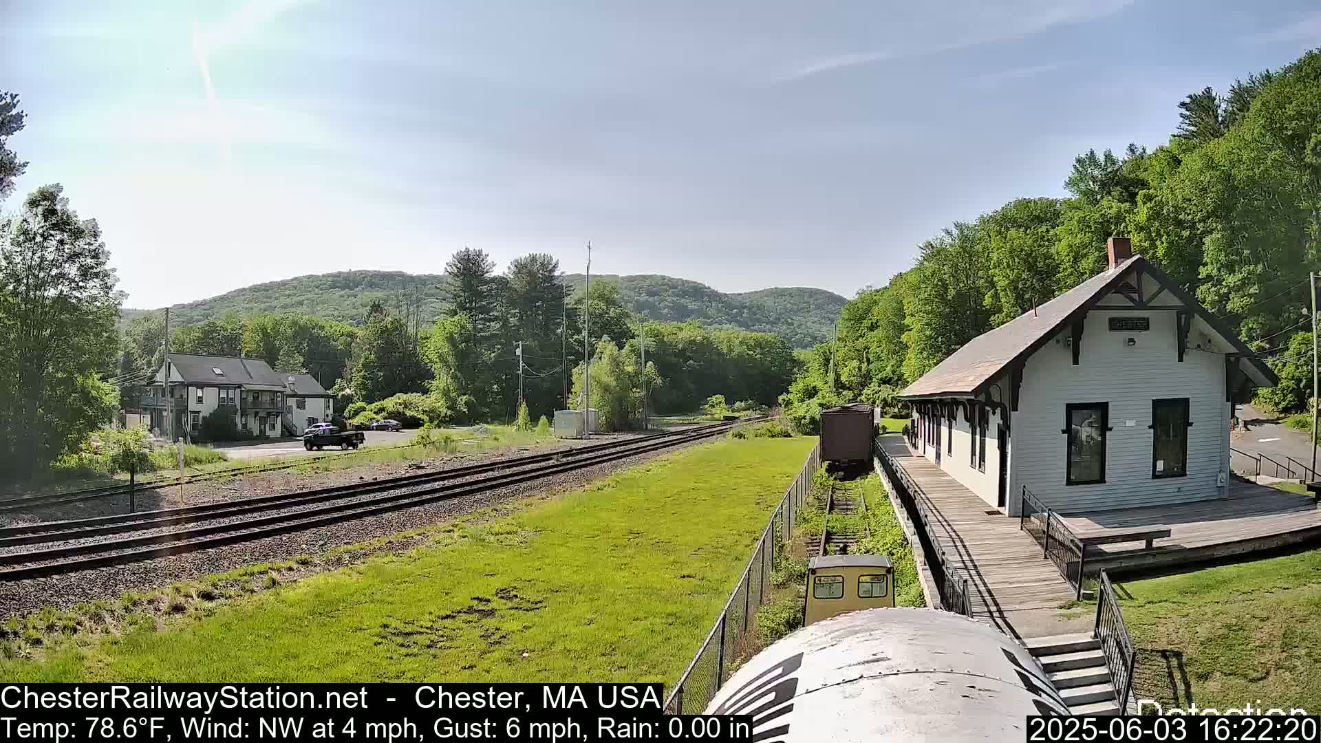 A train approaches a small, white train station on a sunny day with light northwest winds, nestled amongst green hills and trees.