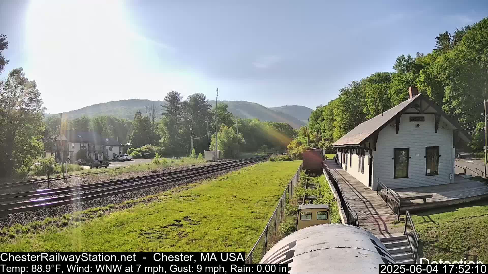 A sunny view of a train station, train tracks, and surrounding greenery on a partly sunny day with a light wind.