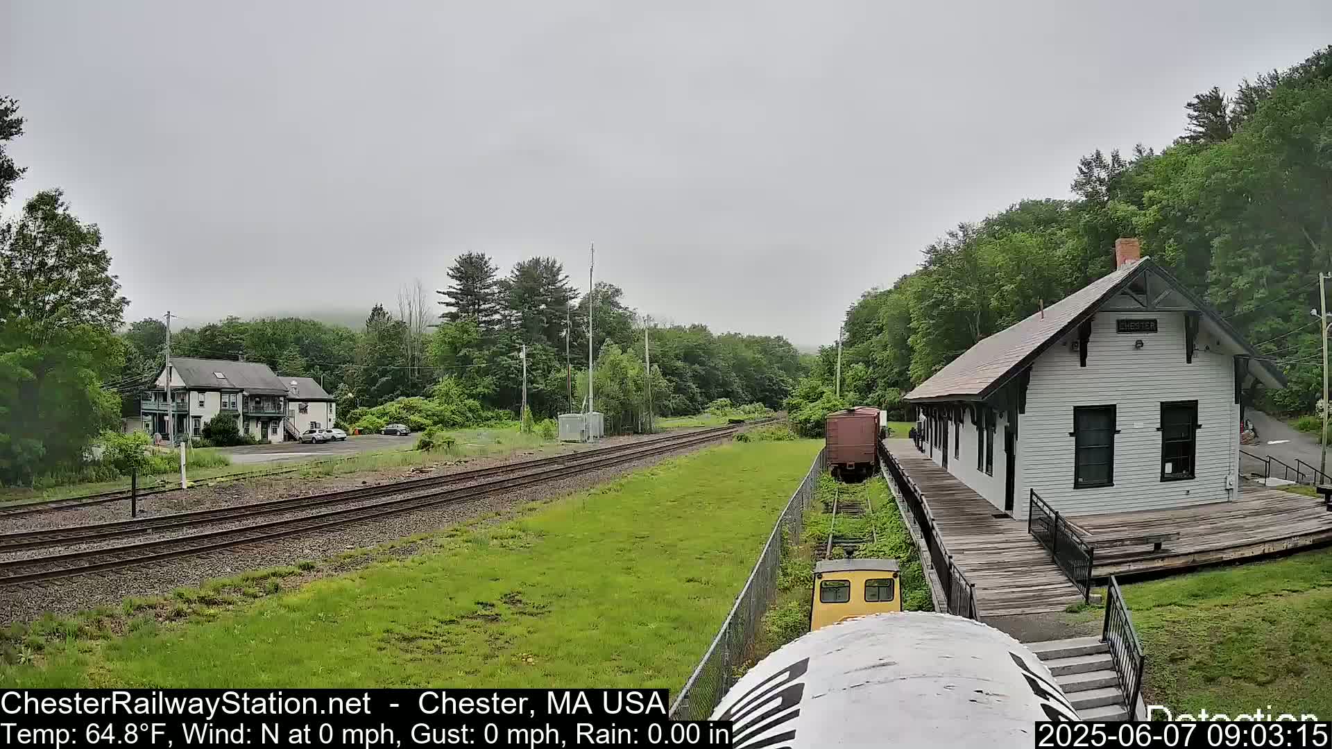 A small, white train station sits beside railroad tracks on a cloudy day, with a small train car and lush green vegetation visible.