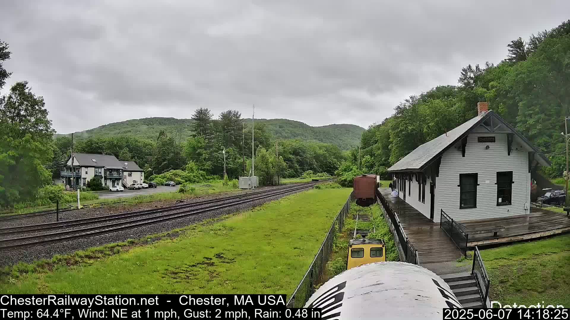 A train approaches a small, white train station situated beside railroad tracks, on a rainy day in a lush green valley.