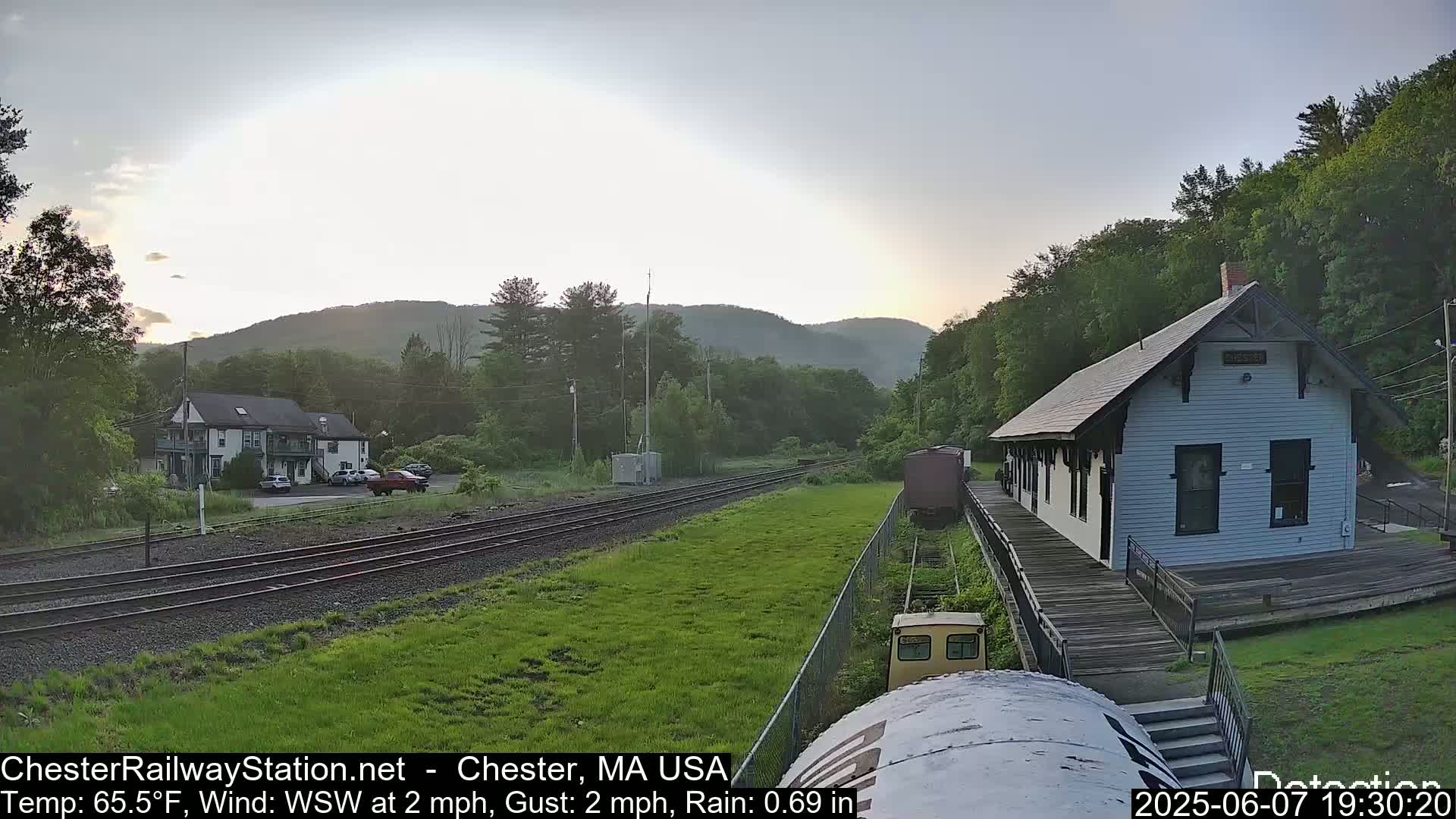 A train station and a residential building are situated on a grassy area beside railroad tracks, with mountains in the background under a partly cloudy, evening sky.