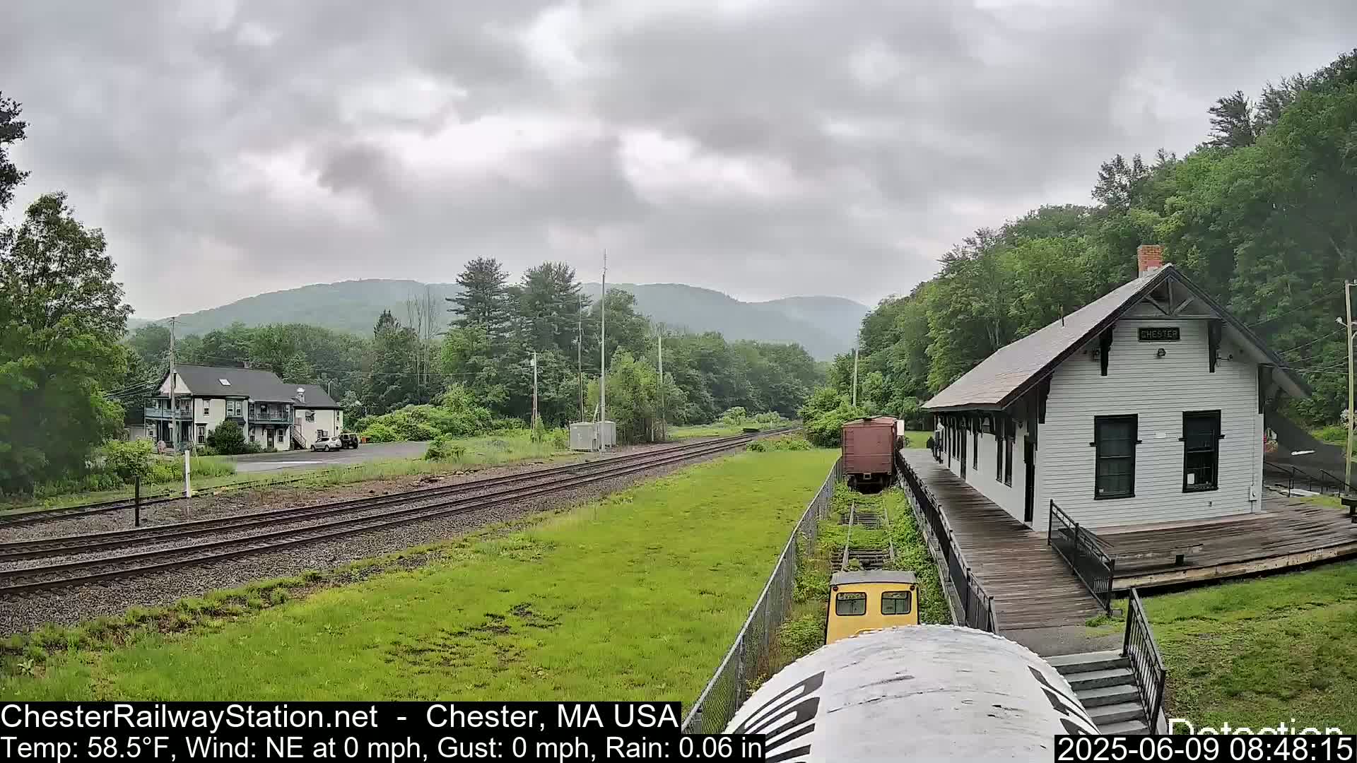 A white train station sits on a wooden platform beside train tracks, with a light rain falling under an overcast sky, and green hills and trees in the background.