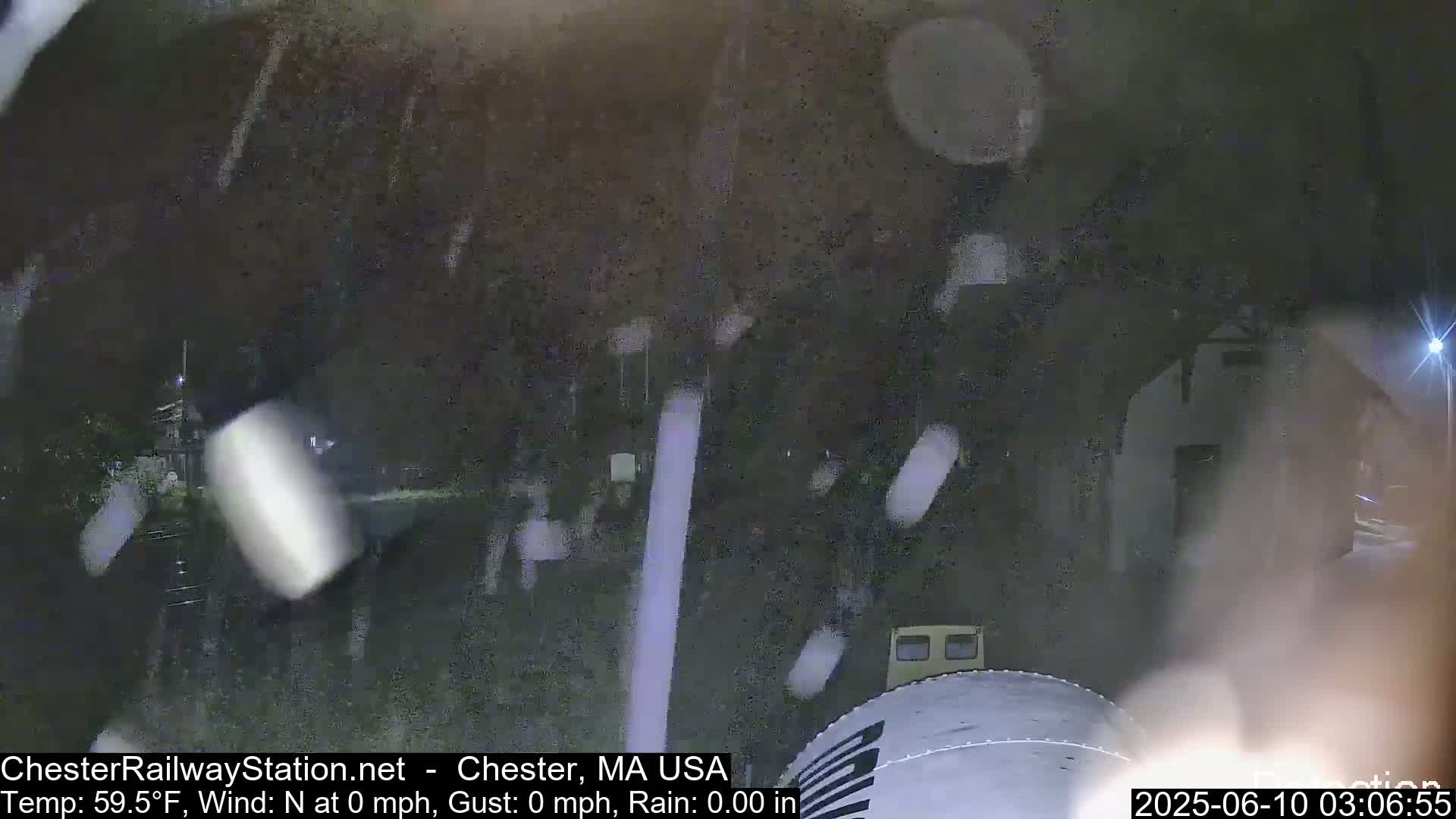 A nighttime view shows rain falling near a train as it moves through a dimly lit area.