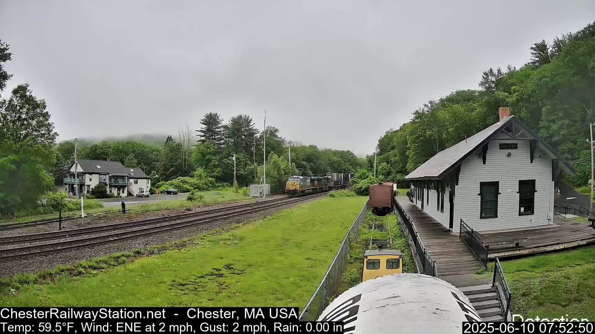 A freight train passes a white train station on an overcast day.