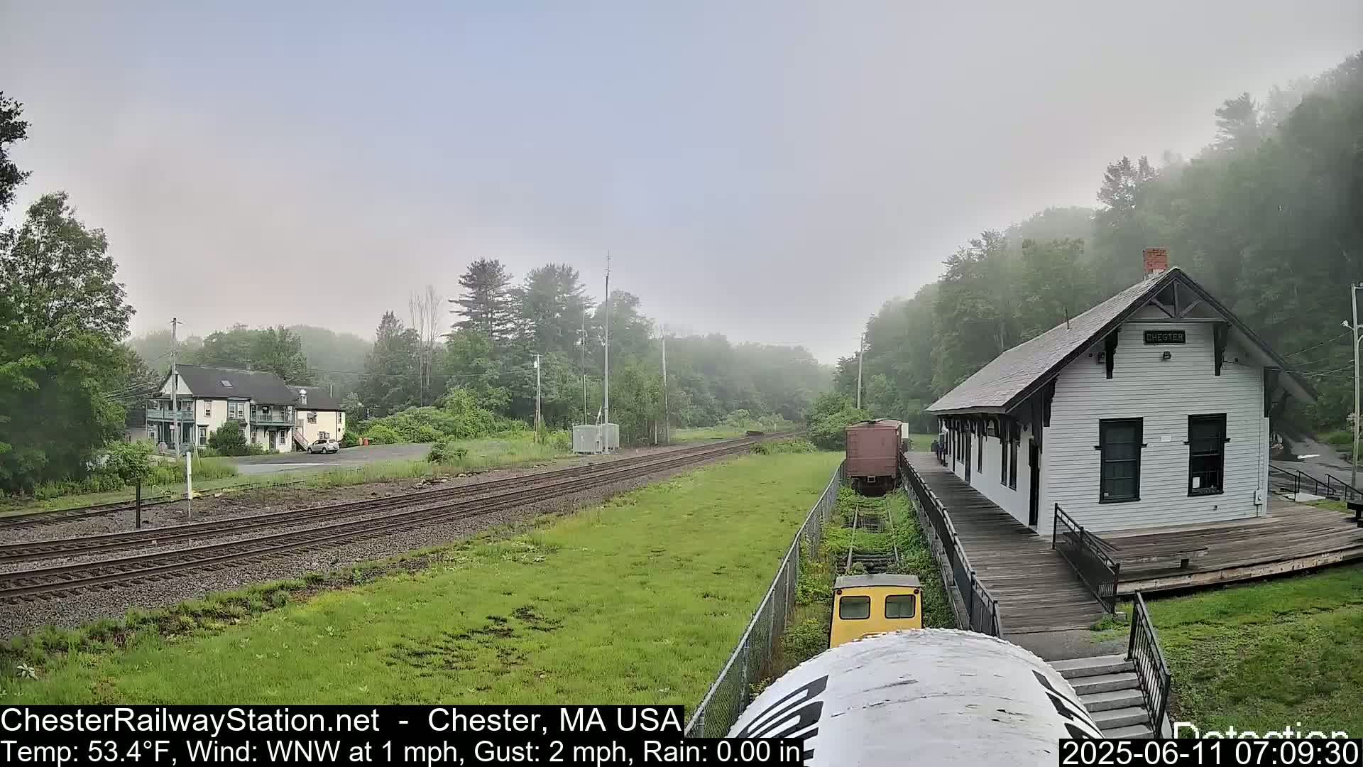 A foggy scene shows a train station and tracks, with a small building and some trees in the background.