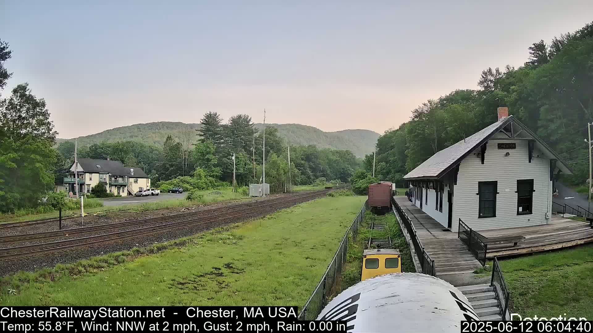 A white train station stands beside a railway track, with a two-story building and green hills in the background under a partly cloudy sky.