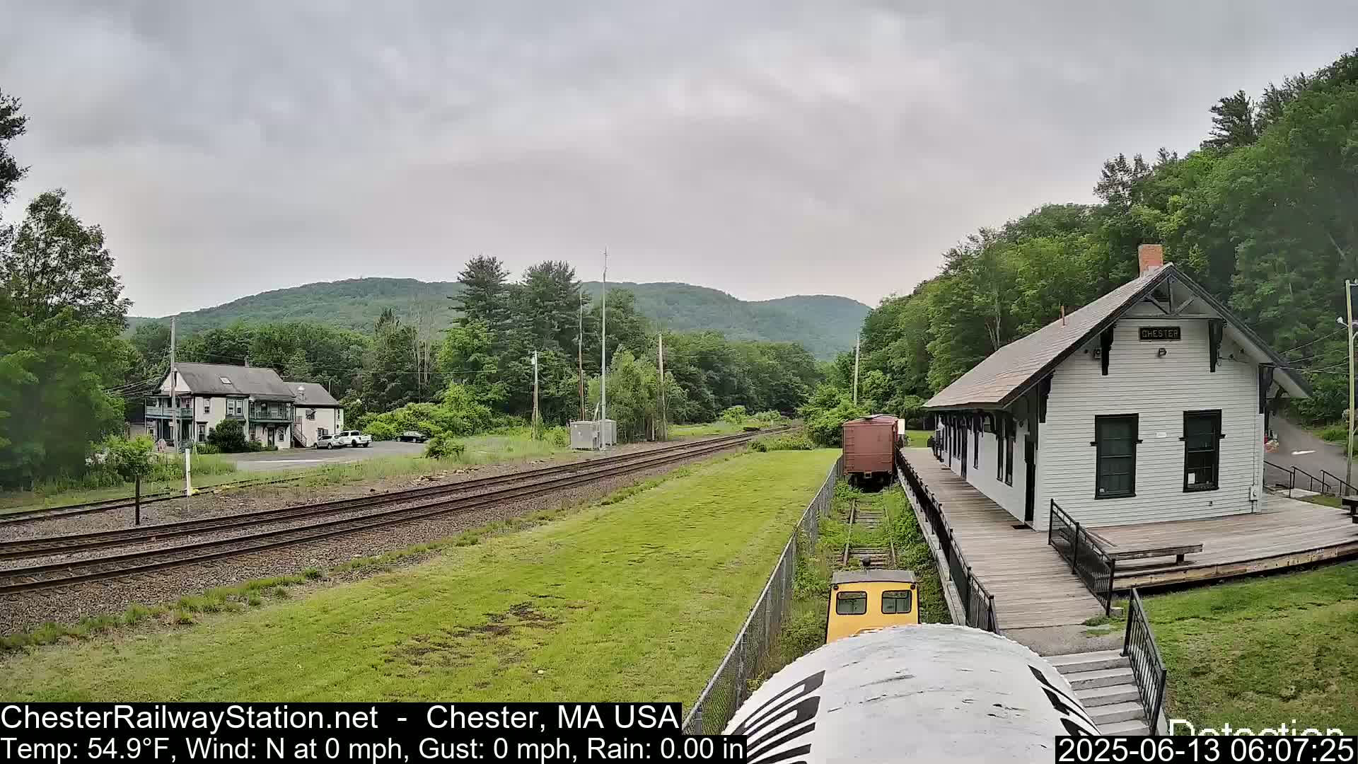 A train approaches a white train station in a verdant valley under an overcast sky.