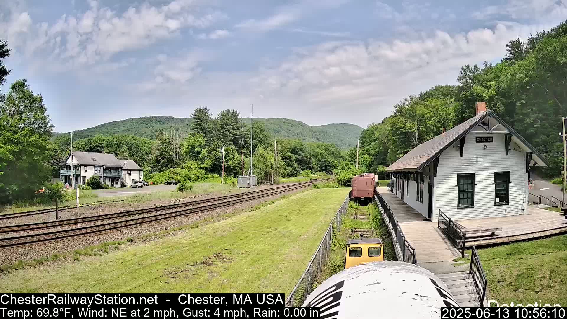 A train approaches a white train station next to a two-story building, all situated in a green, hilly landscape under a partly cloudy sky.