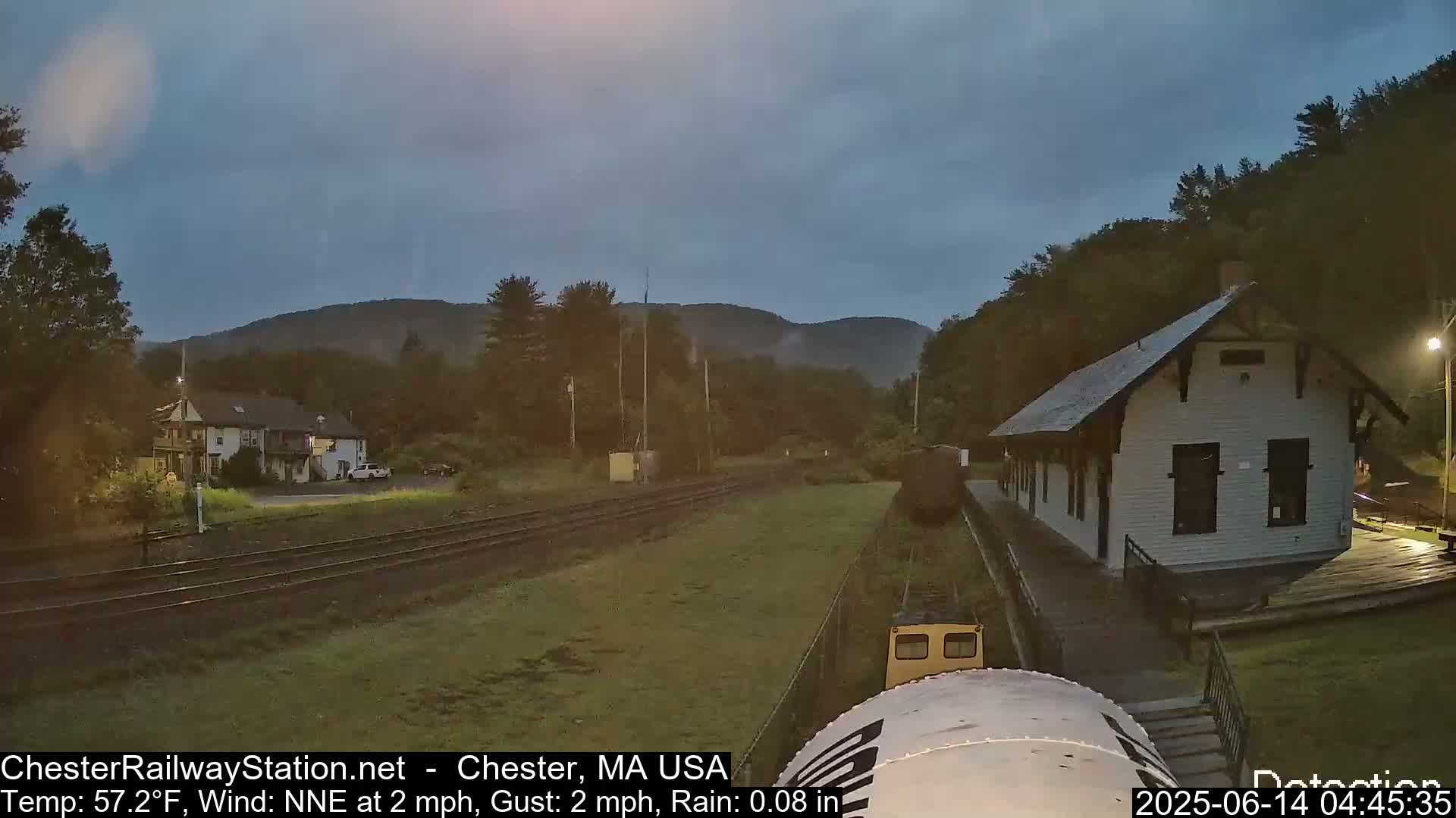 A train car sits on railway tracks overlooking a small station and a building next to a hill under a lightly rainy, overcast sky.