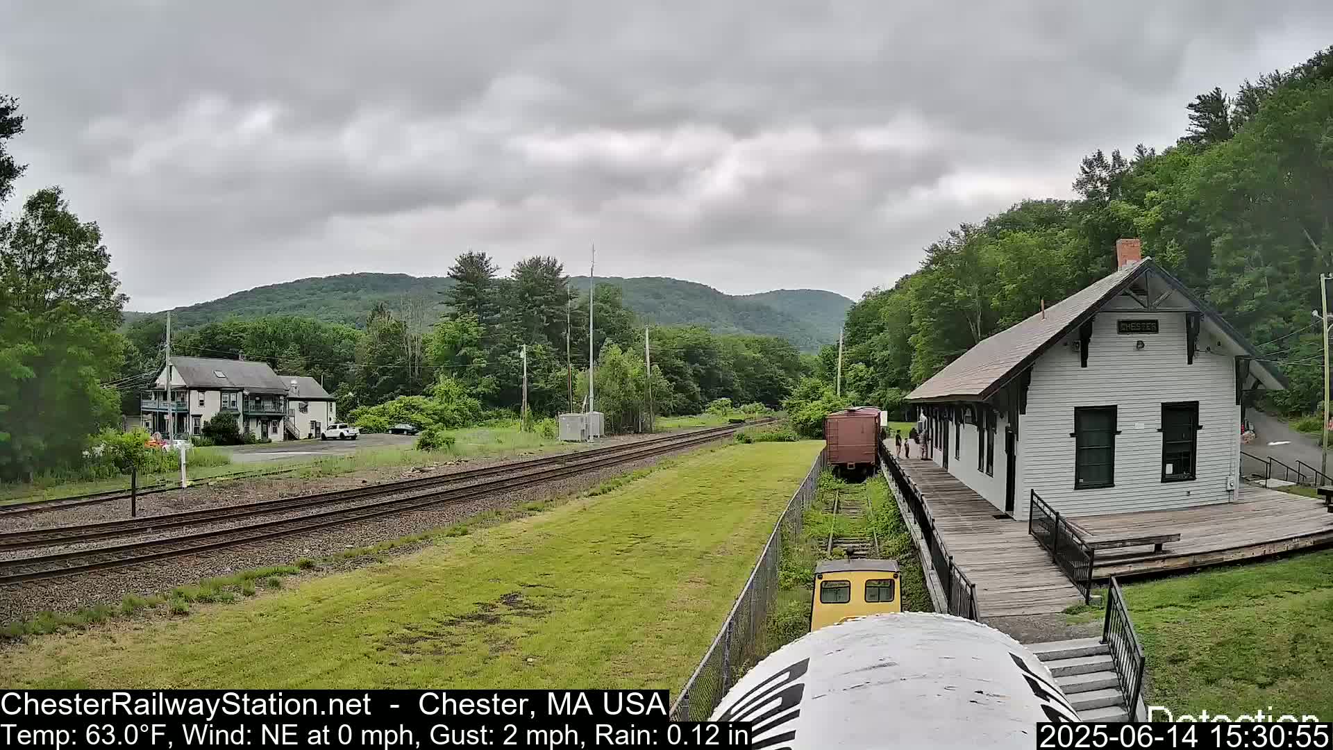 A train car sits on railway tracks overlooking a small station and a building next to a hill under a lightly rainy, overcast sky.