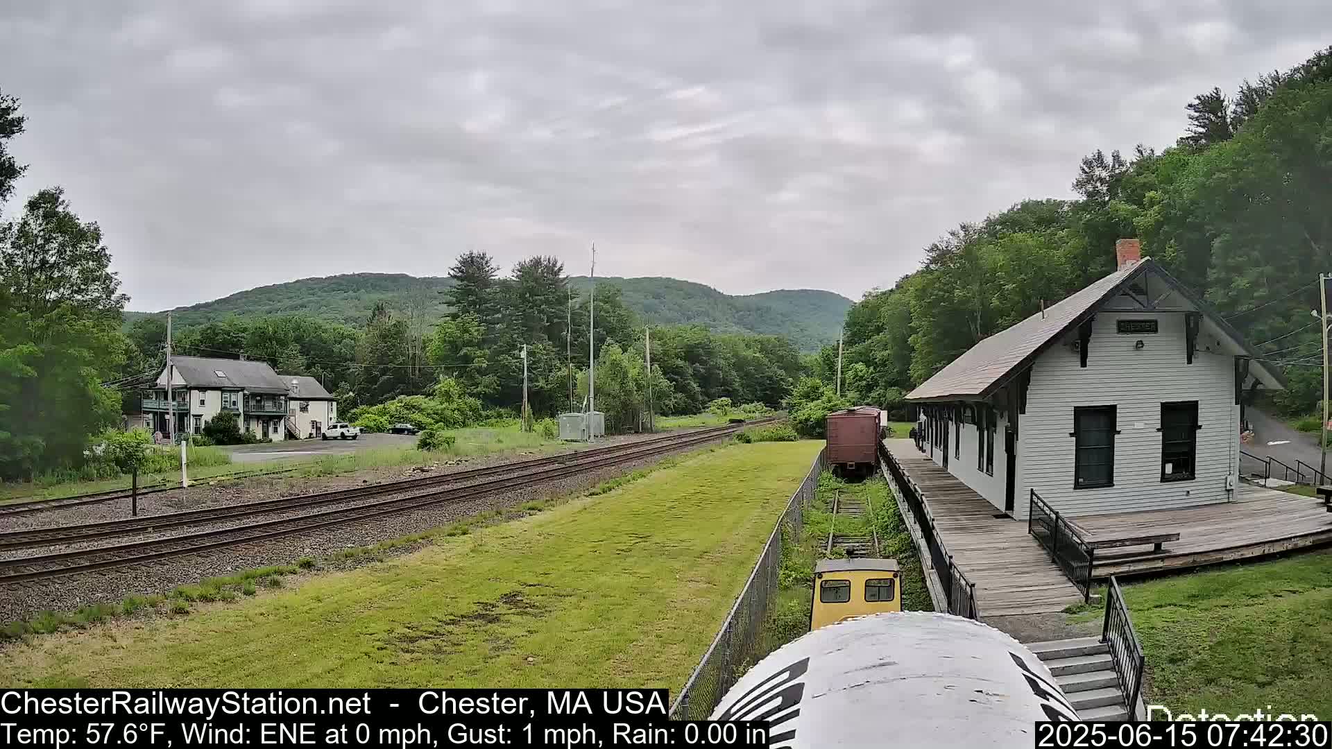 A train is approaching a white train station on a slightly overcast day, with green hills and trees in the background.