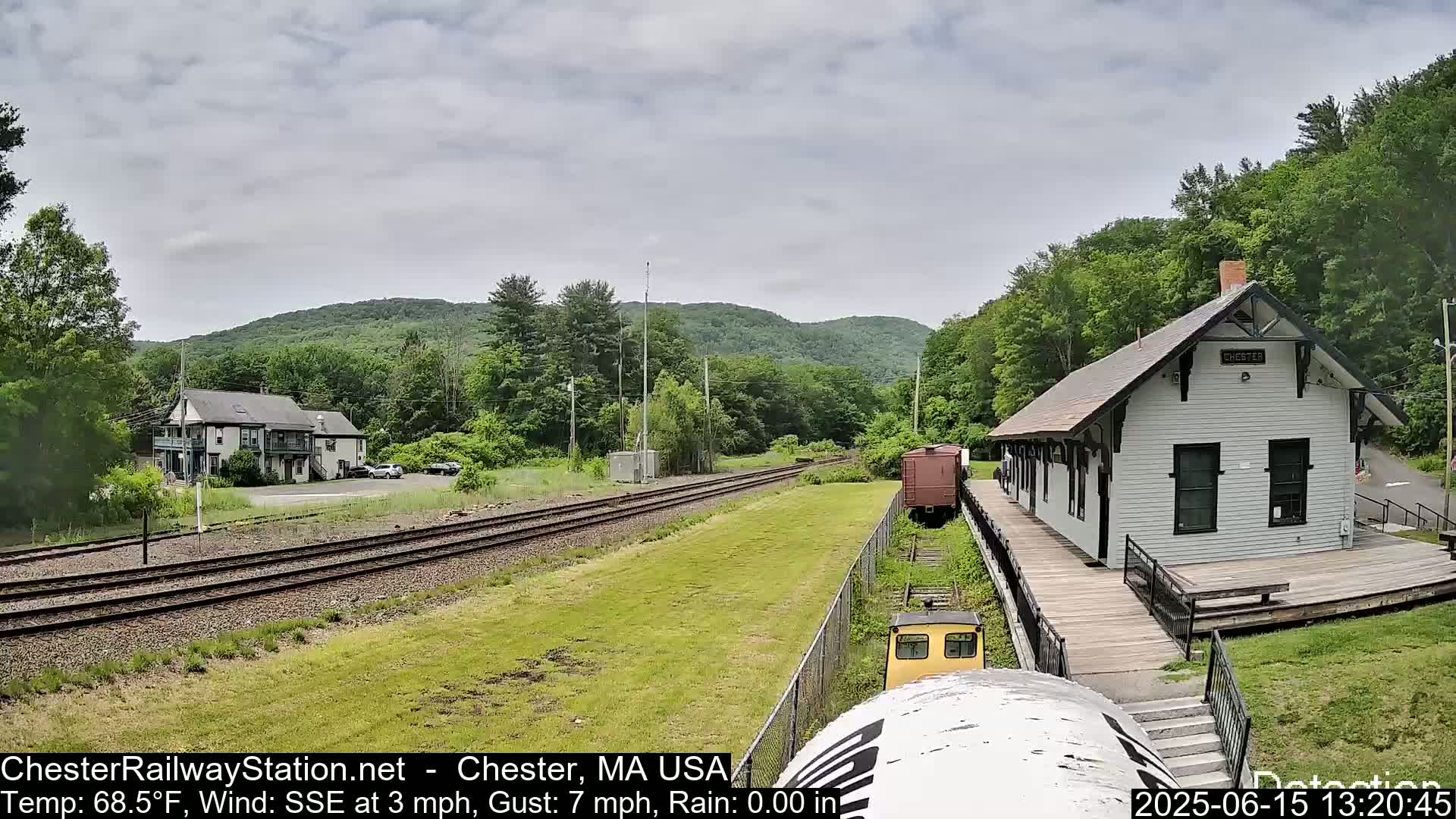 A train approaches a small, white train station nestled in a green, hilly landscape under a partly cloudy sky.