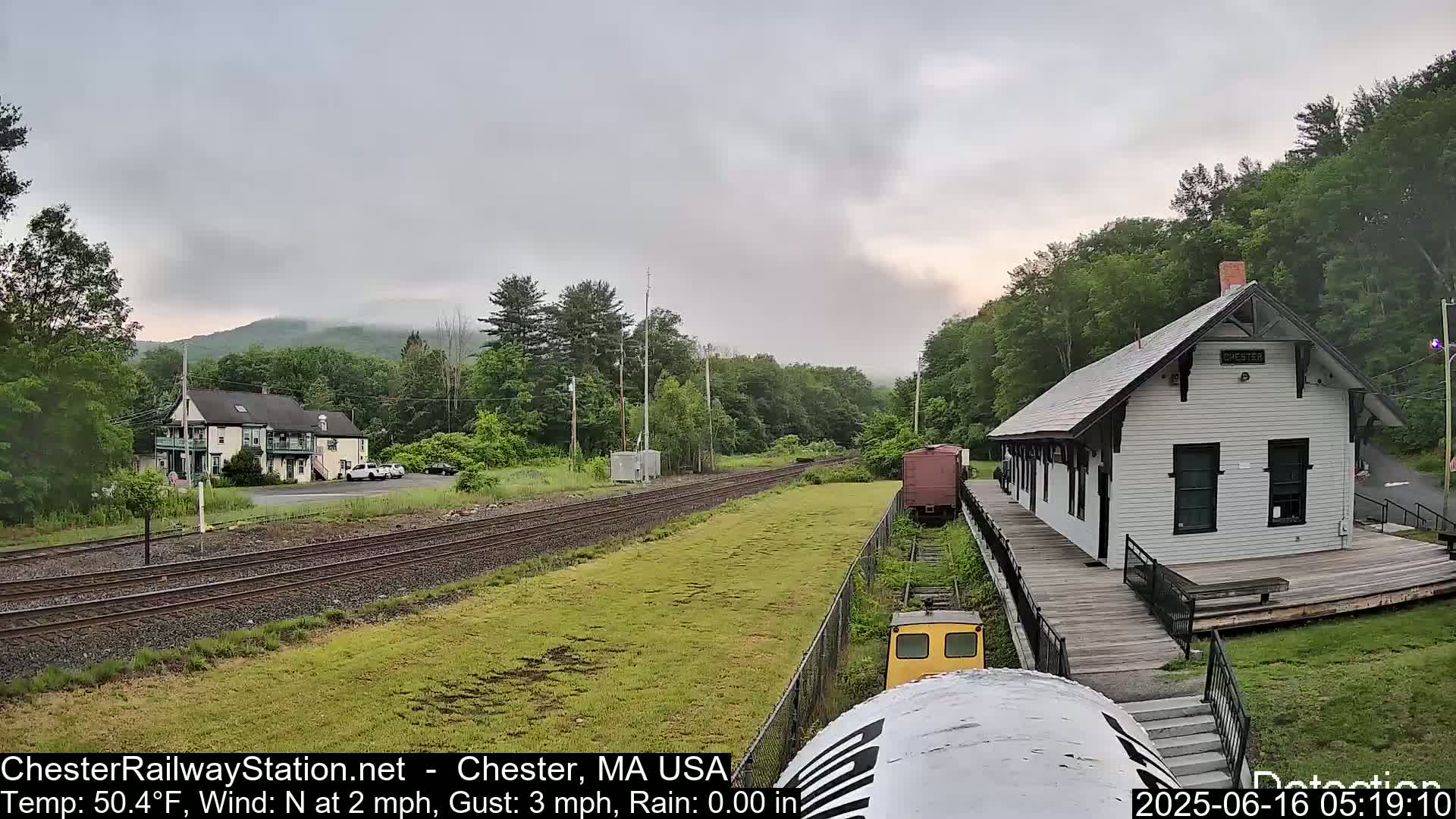 A white train station sits beside a railway track under a partly cloudy sky, with a small town and green hills in the background.